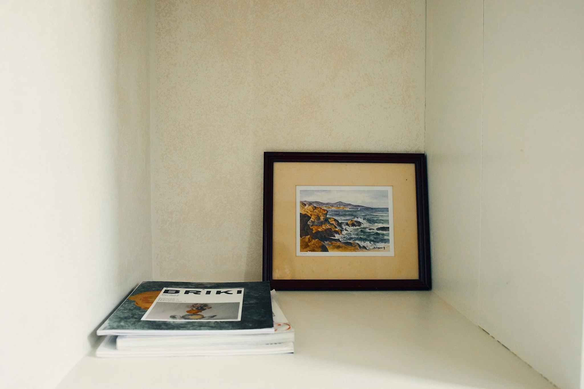 A corner of a room with a framed landscape painting of rocky cliffs and ocean waves hanging on the wall. In the foreground on a white shelf, there are newspapers and a magazine, one titled 'BRIK'.