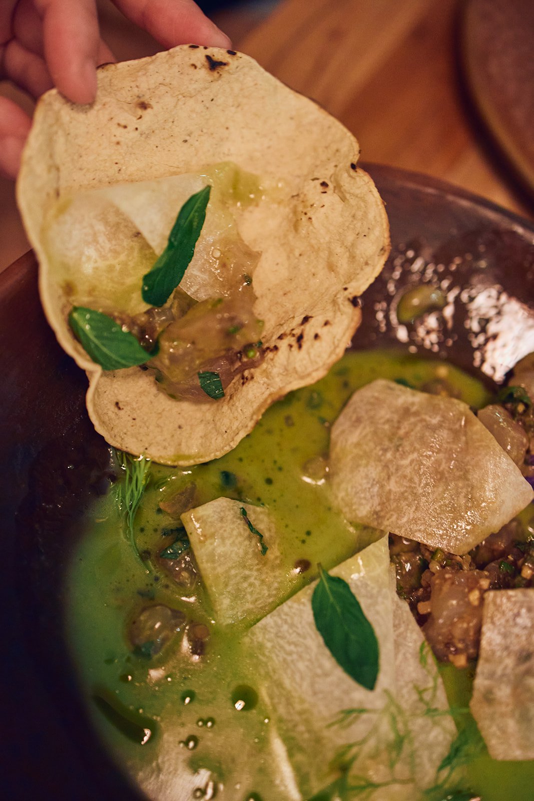 Tortilla chip dipped in green sauce with herbs, onions, and seeds, in a bowl of green sauce with herbs and onion pieces.