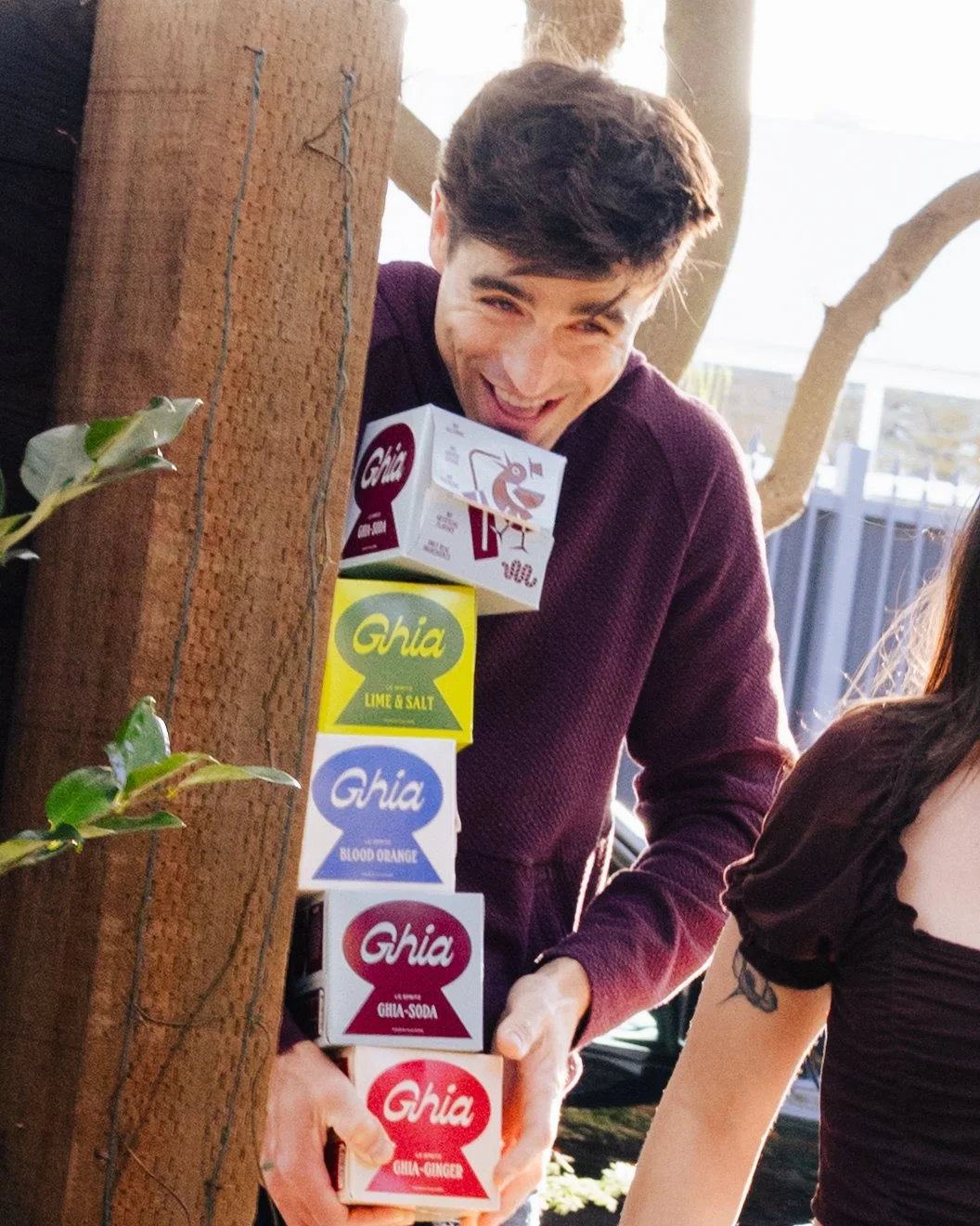 A smiling young man peeking from behind a tree, holding stacked boxes of Ghia soda with flavored labels, outdoors with trees and a fence in the background.
