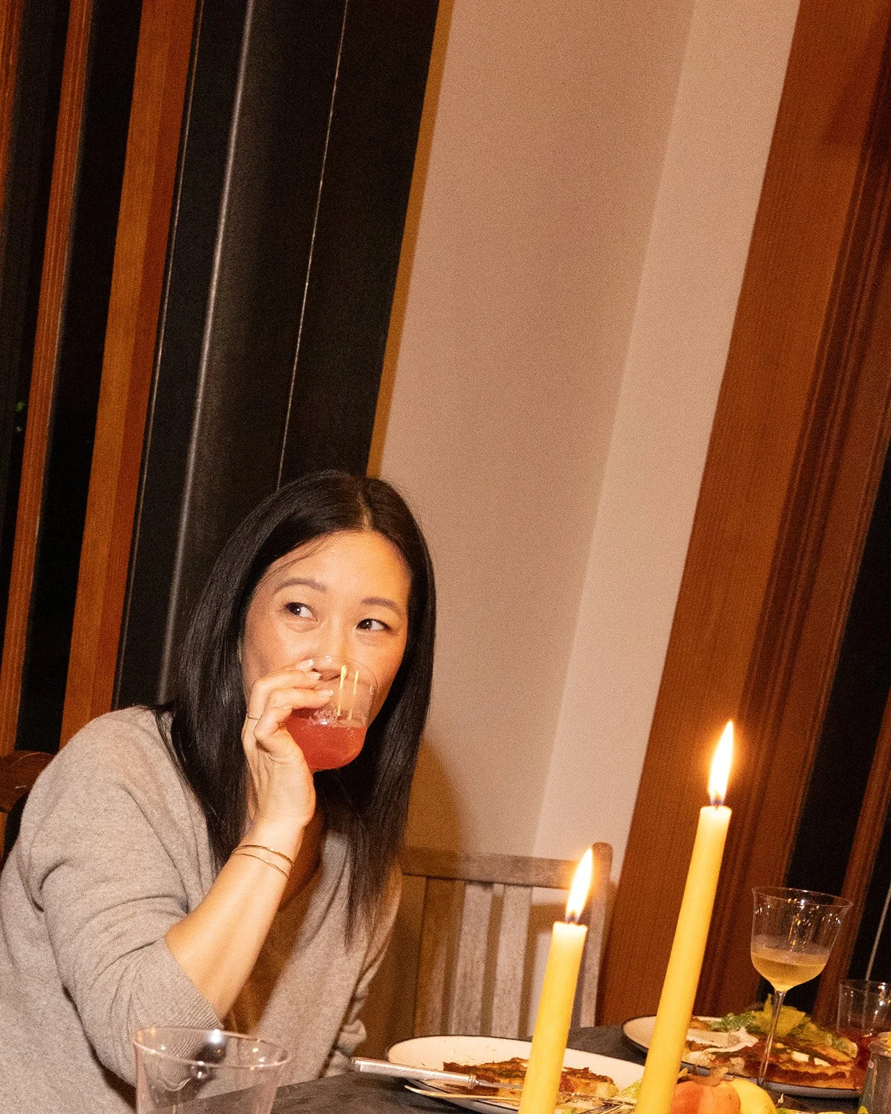 A woman with black hair and a beige sweater sitting at a dinner table, sipping a red drink from a glass, with lit candles and pizza on the table.