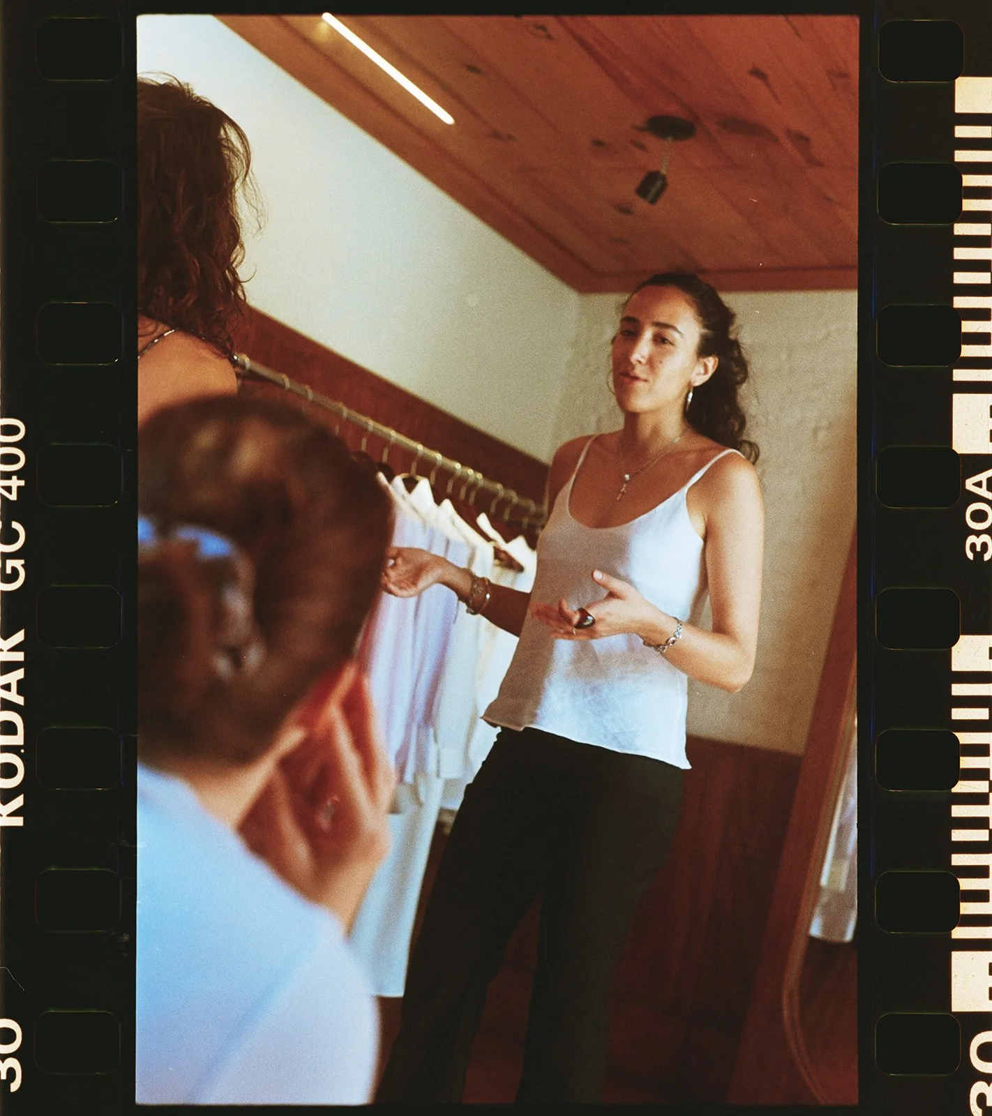 A woman with dark curly hair wearing a white tank top and black pants standing indoors, speaking to a group of people, with a clothing rack and wooden ceiling visible in the background.