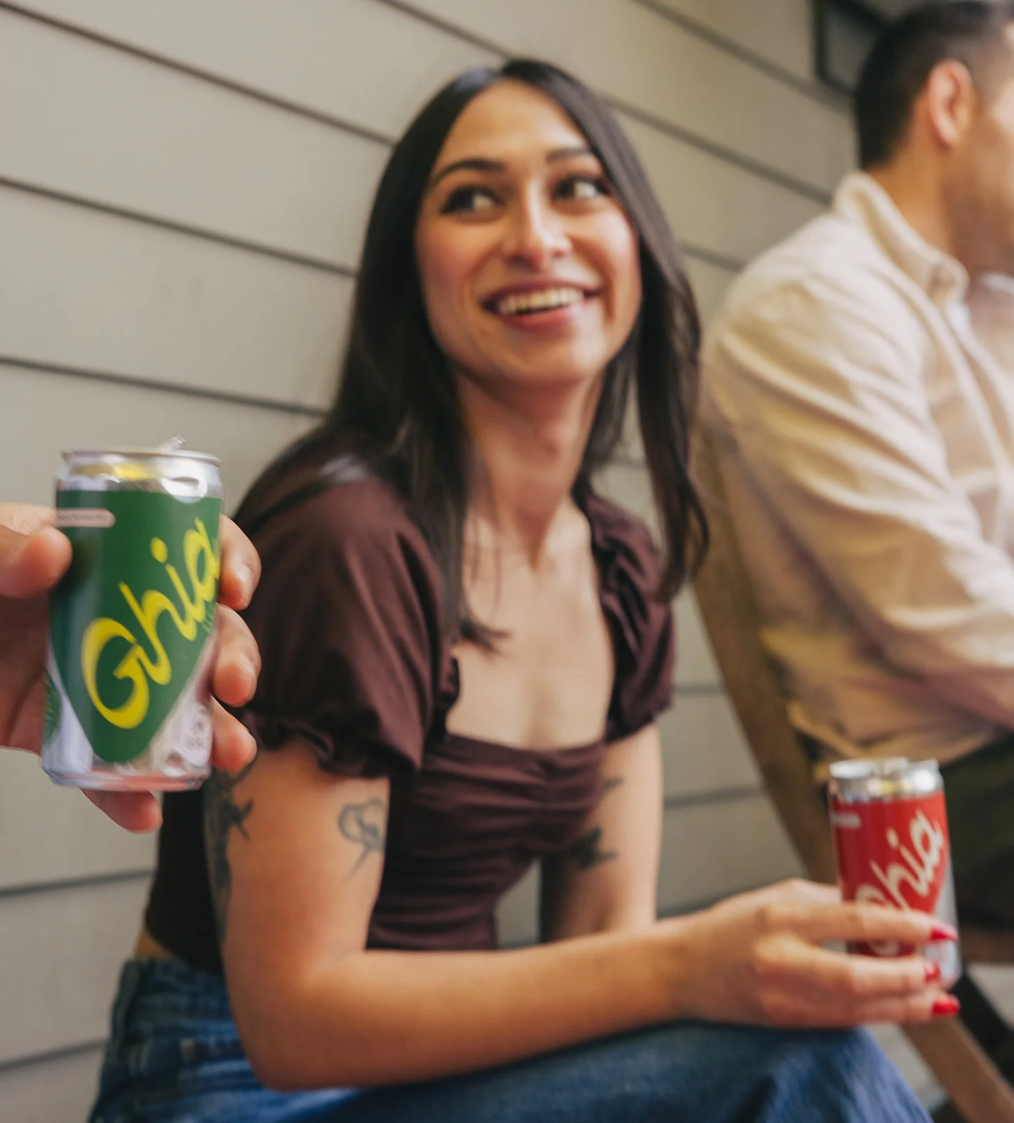 A woman smiling and sitting with a man beside her, both holding Ghia soda cans, in a casual setting against a wooden wall.