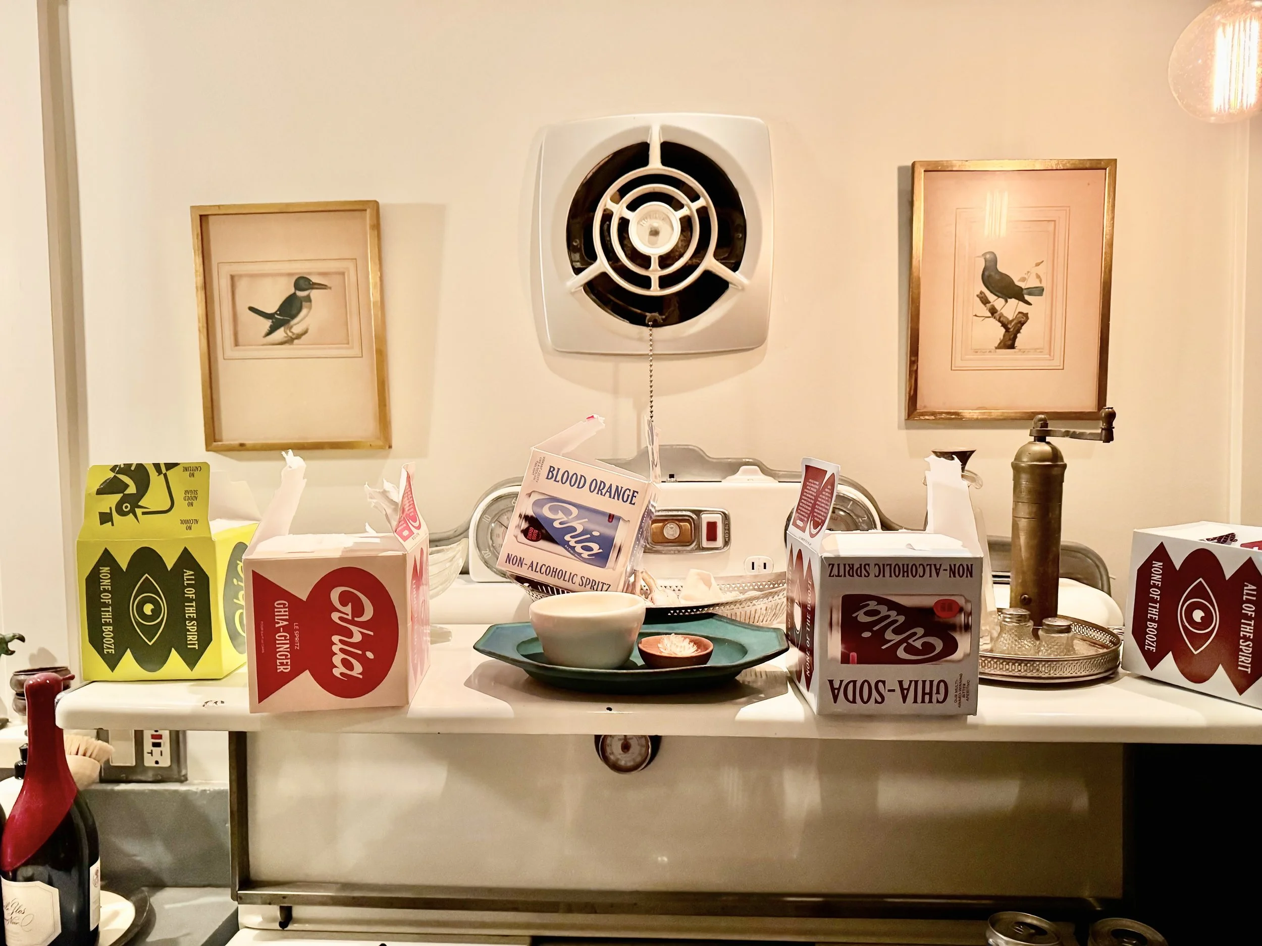 A cluttered white table with juice boxes, a bowl, and a small dish in front of a wall with framed bird pictures. Above the table there's a white vent.
