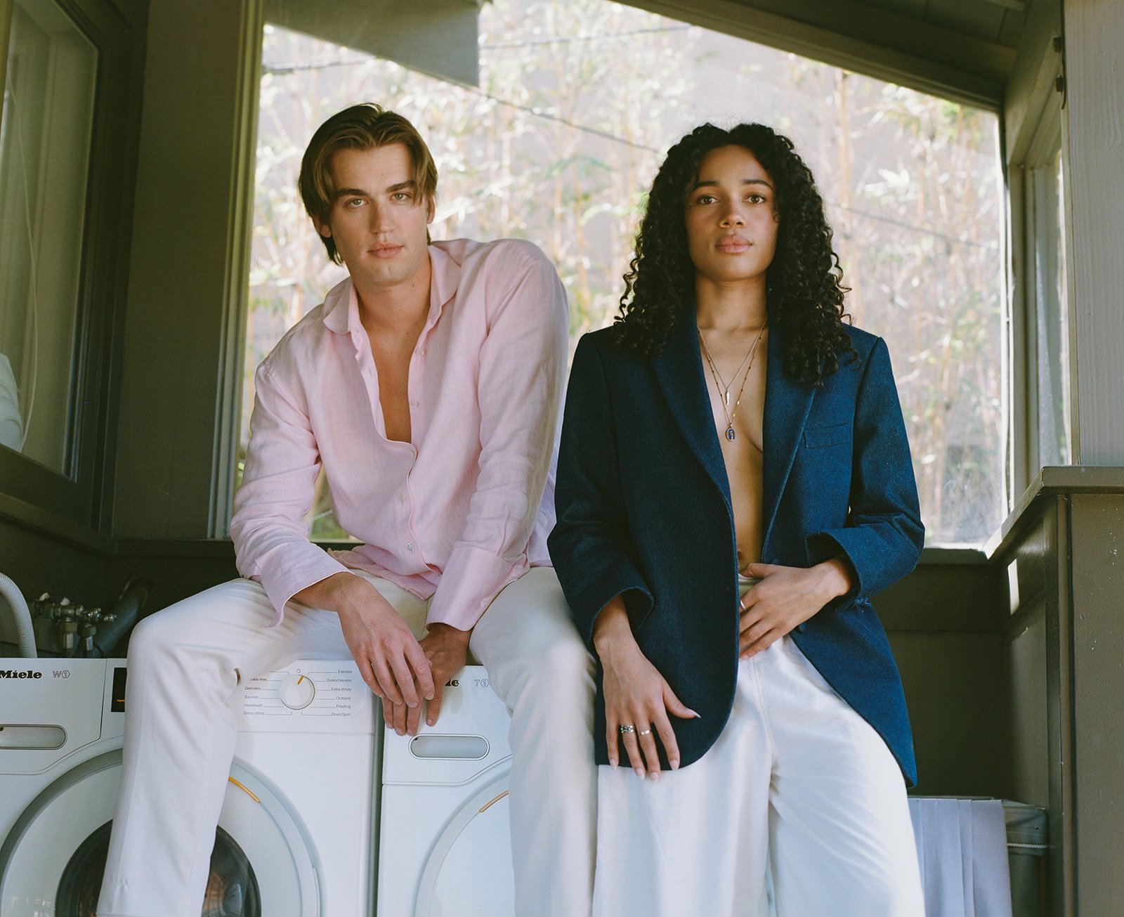 A man and woman sitting on a washing machine in a laundry room with large windows and natural light. The man is wearing a light pink shirt and white pants, and the woman is wearing a dark blue blazer over a top and white pants.