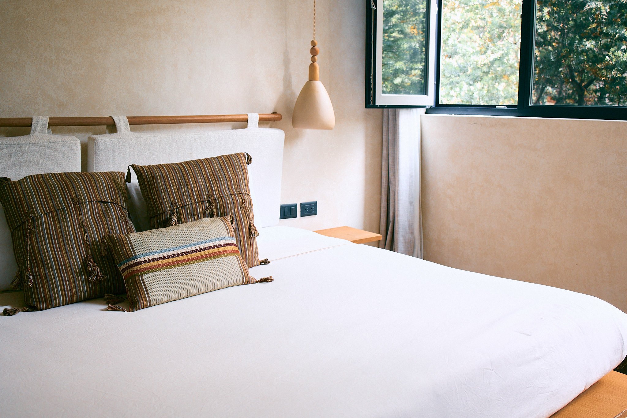 A bedroom with a bed, four patterned pillows, an open window showing trees outside, a hanging lamp, and two electrical outlets on the wall.