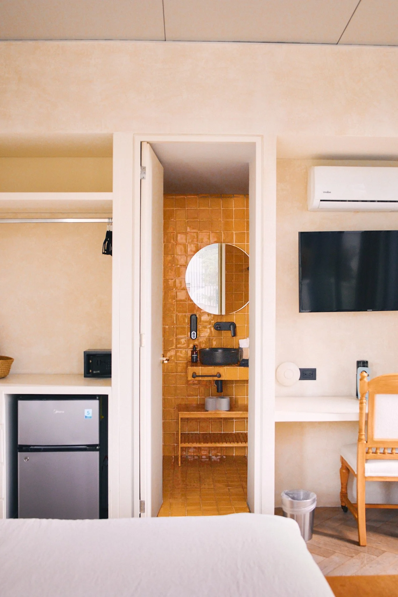 Living room with beige walls, a corner with a small refrigerator and black microwave, an air conditioning unit on the wall, a flat-screen TV, wooden chair, and small trash can.