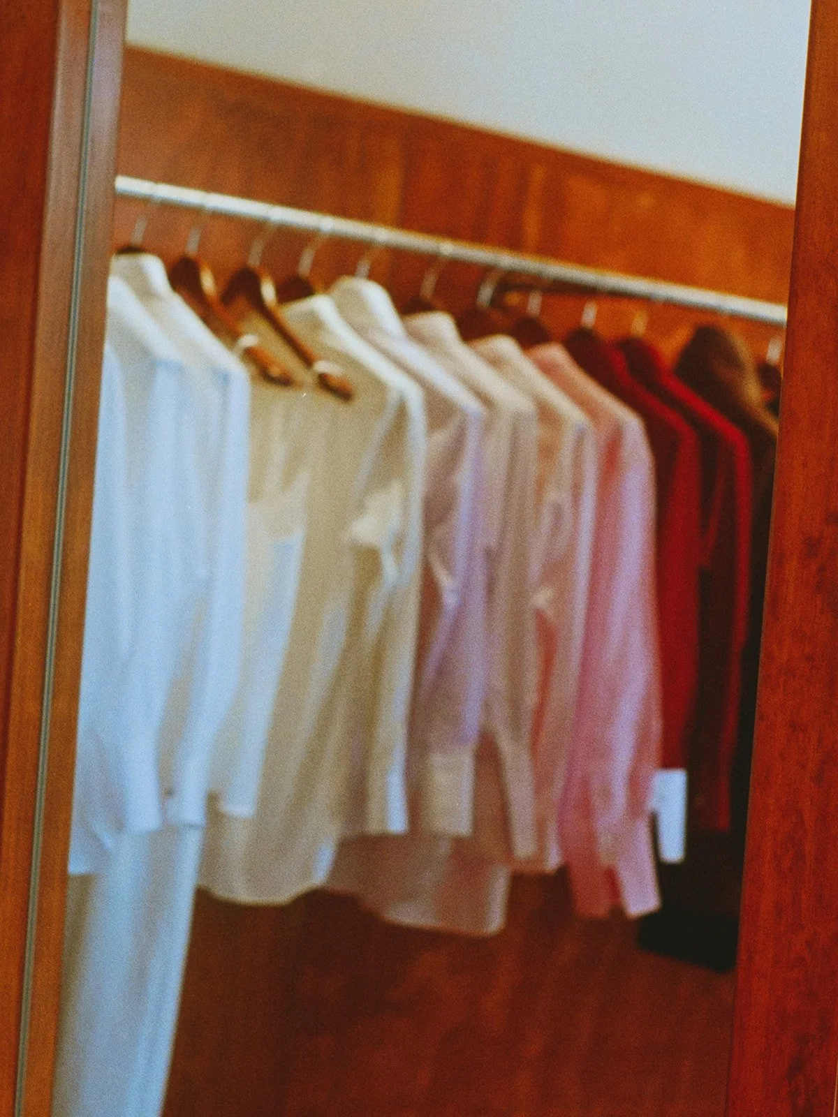 Closet with white, pink, and red shirts hanging on hangers on a wooden rod.