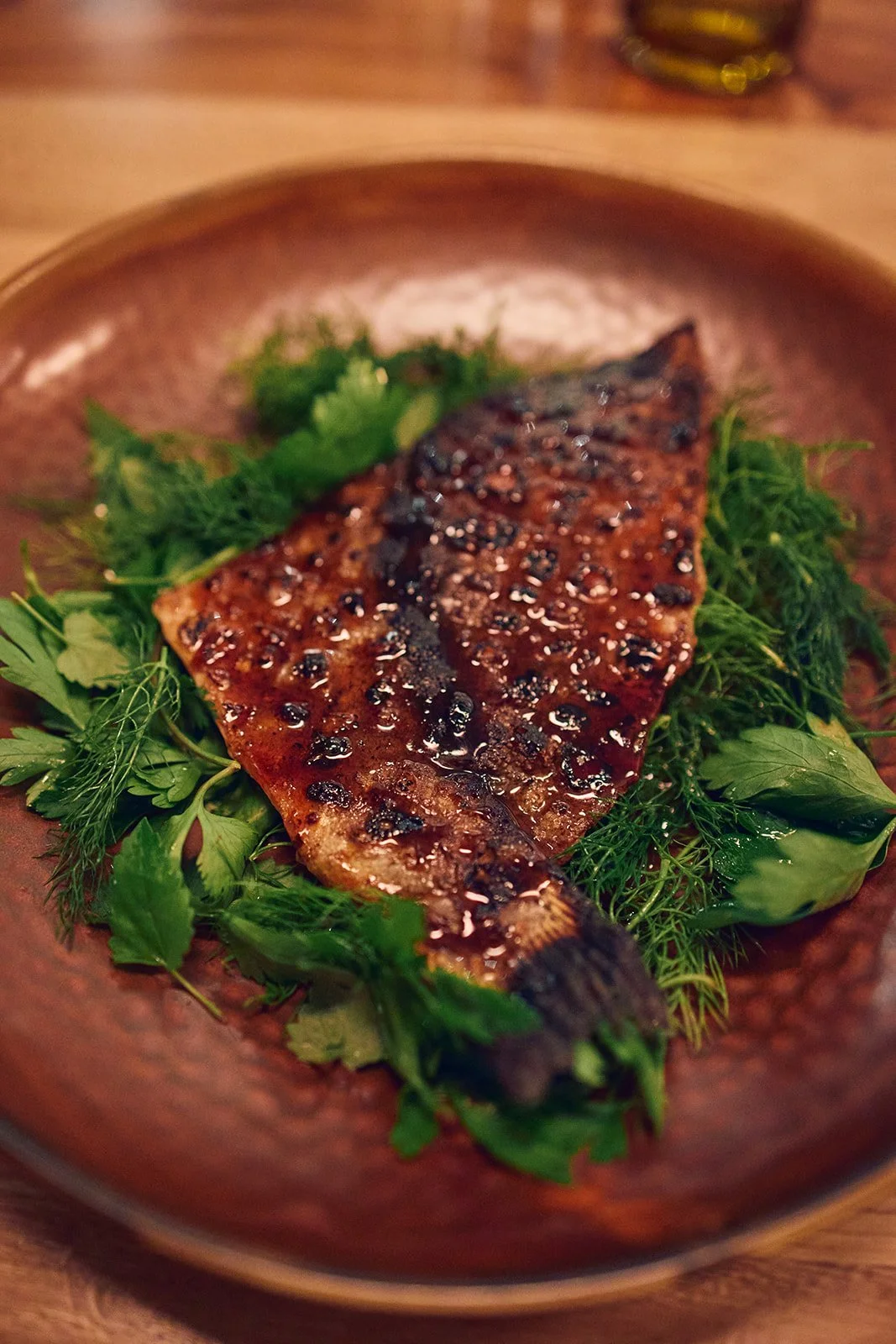Grilled fish fillet with barbecue sauce, served on a bed of fresh green herbs on a brown plate.