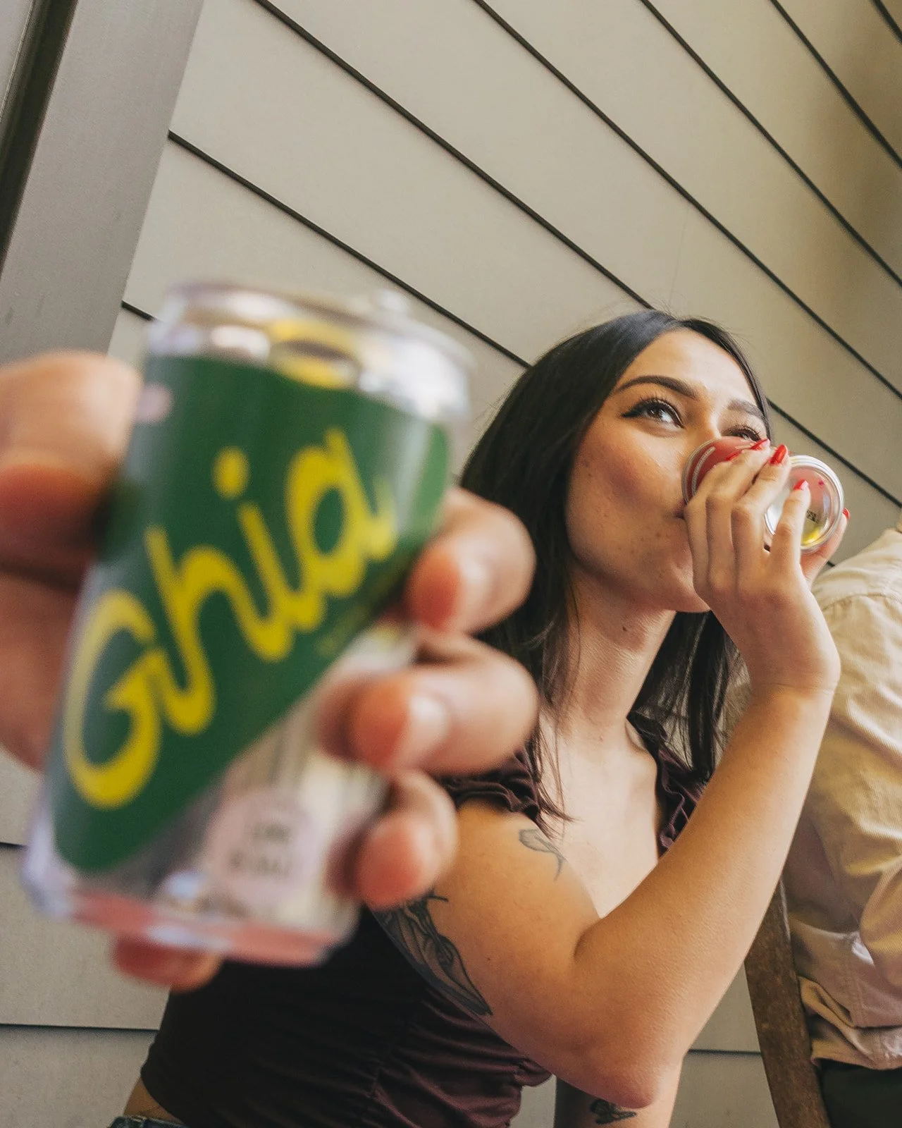 A woman with long dark hair and tattoos on her arm drinks from a small cup while holding a can of Guida soda, with the can blurred in the foreground.