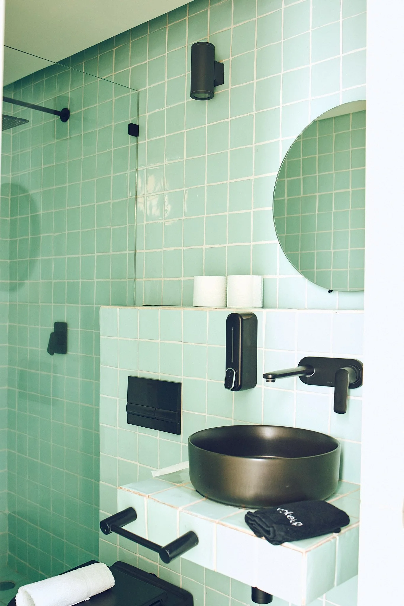 Bathroom with green tiled wall, mirror, black round vessel sink, black faucet, black wall-mounted accessories, toilet paper rolls, and black hand towel on a white tiled counter.