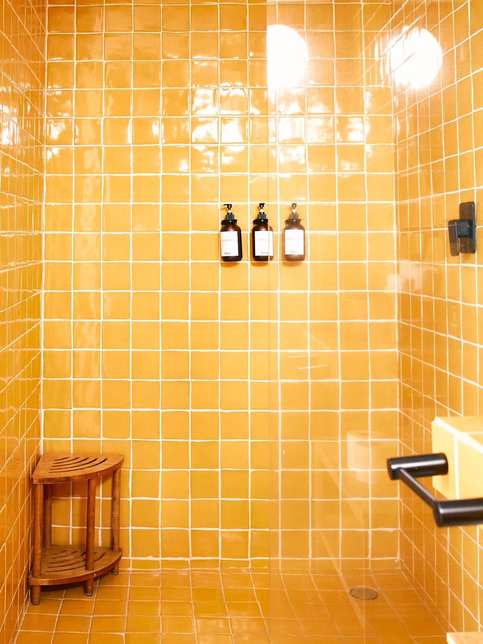 A shower area with orange tiles on the walls and floor, three brown bottles hanging on the wall, a wooden stool in the corner, and a metal handle on the door.