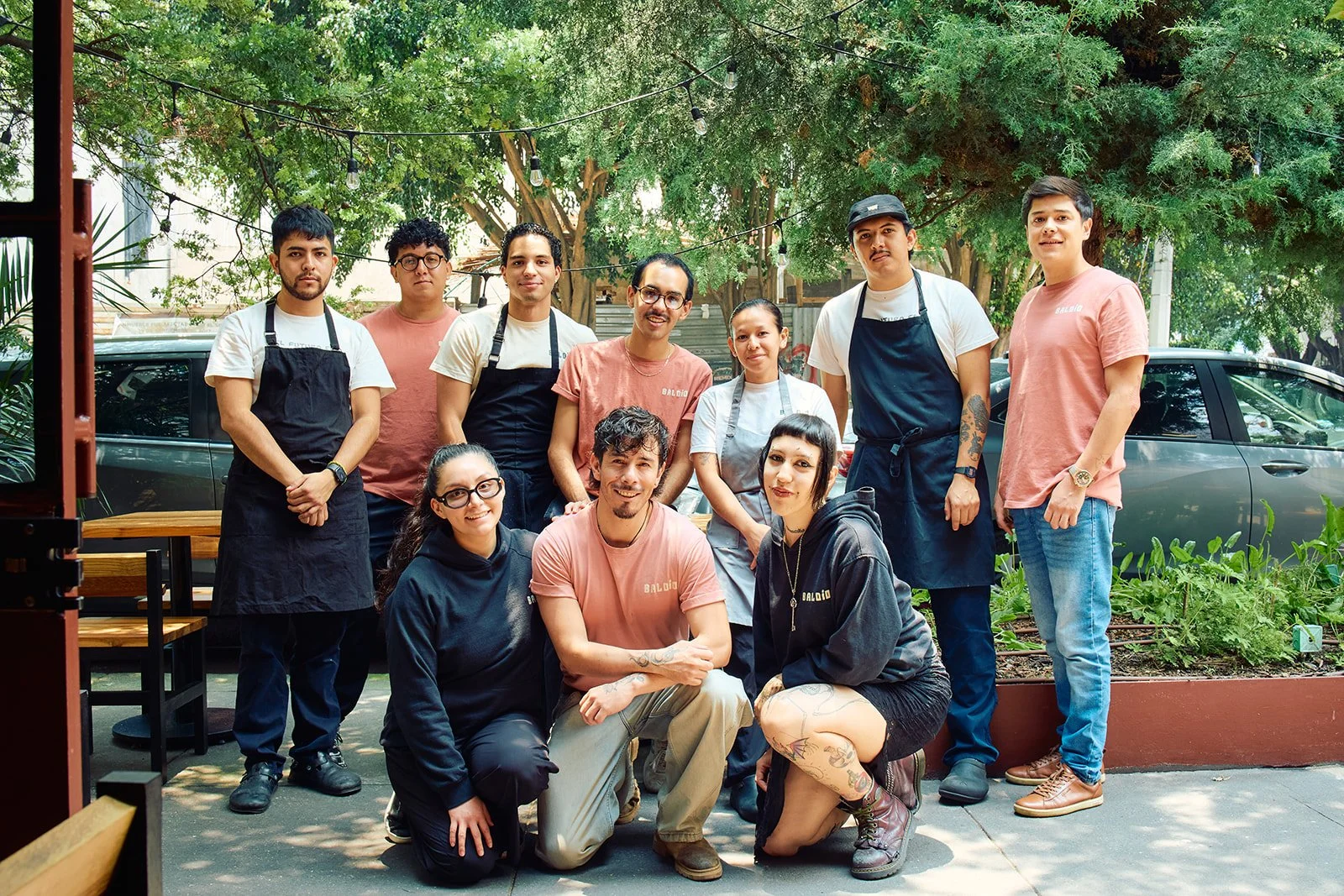 A group of ten people, including restaurant staff and possibly friends or family, posed together outdoors in a garden or patio setting, with trees, greenery, parked cars, and string lights in the background.