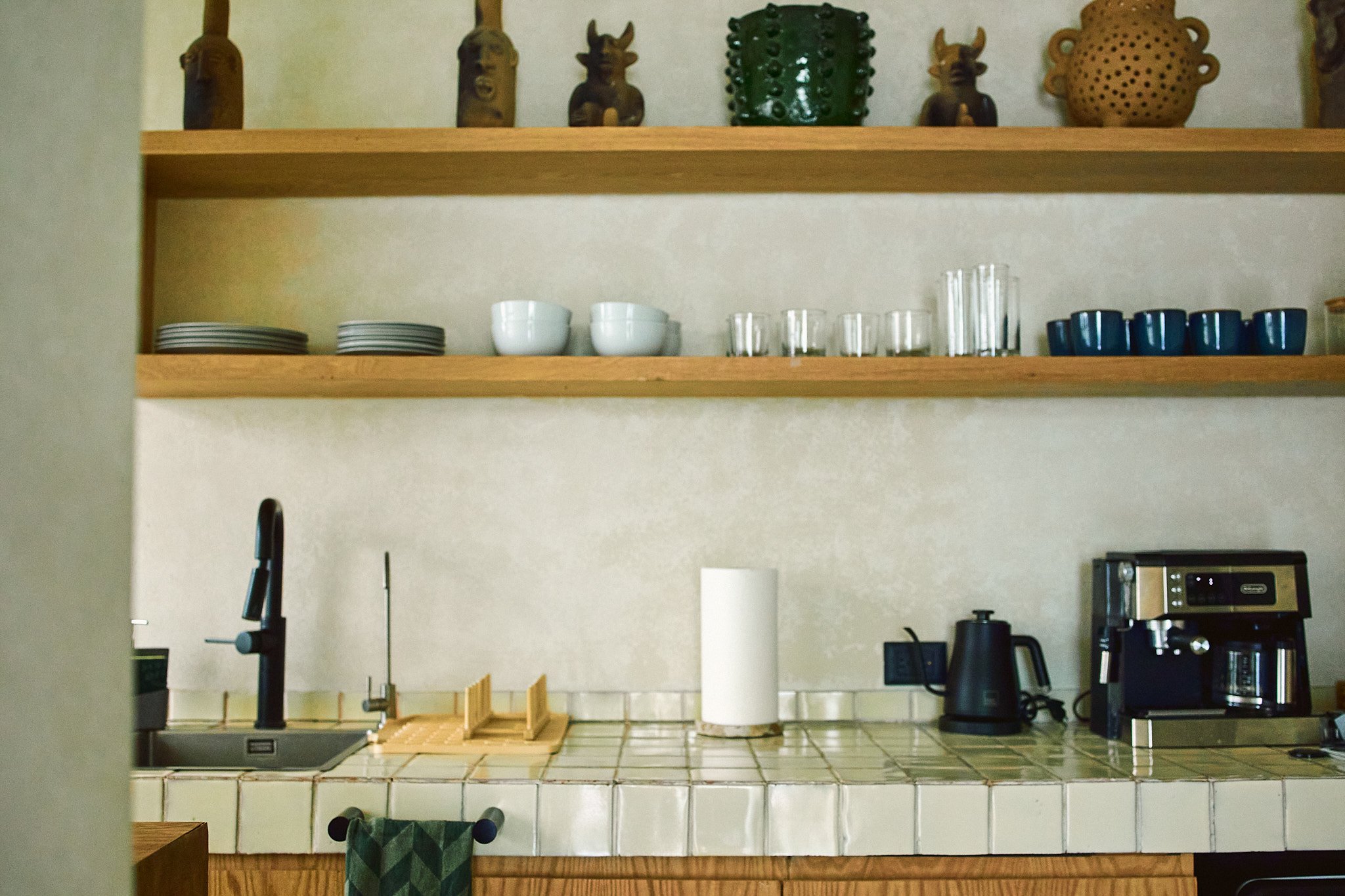 Kitchen shelf with decorative African masks and pottery on top, dishes and glasses on the middle shelf, and various kitchen appliances on the counter below.