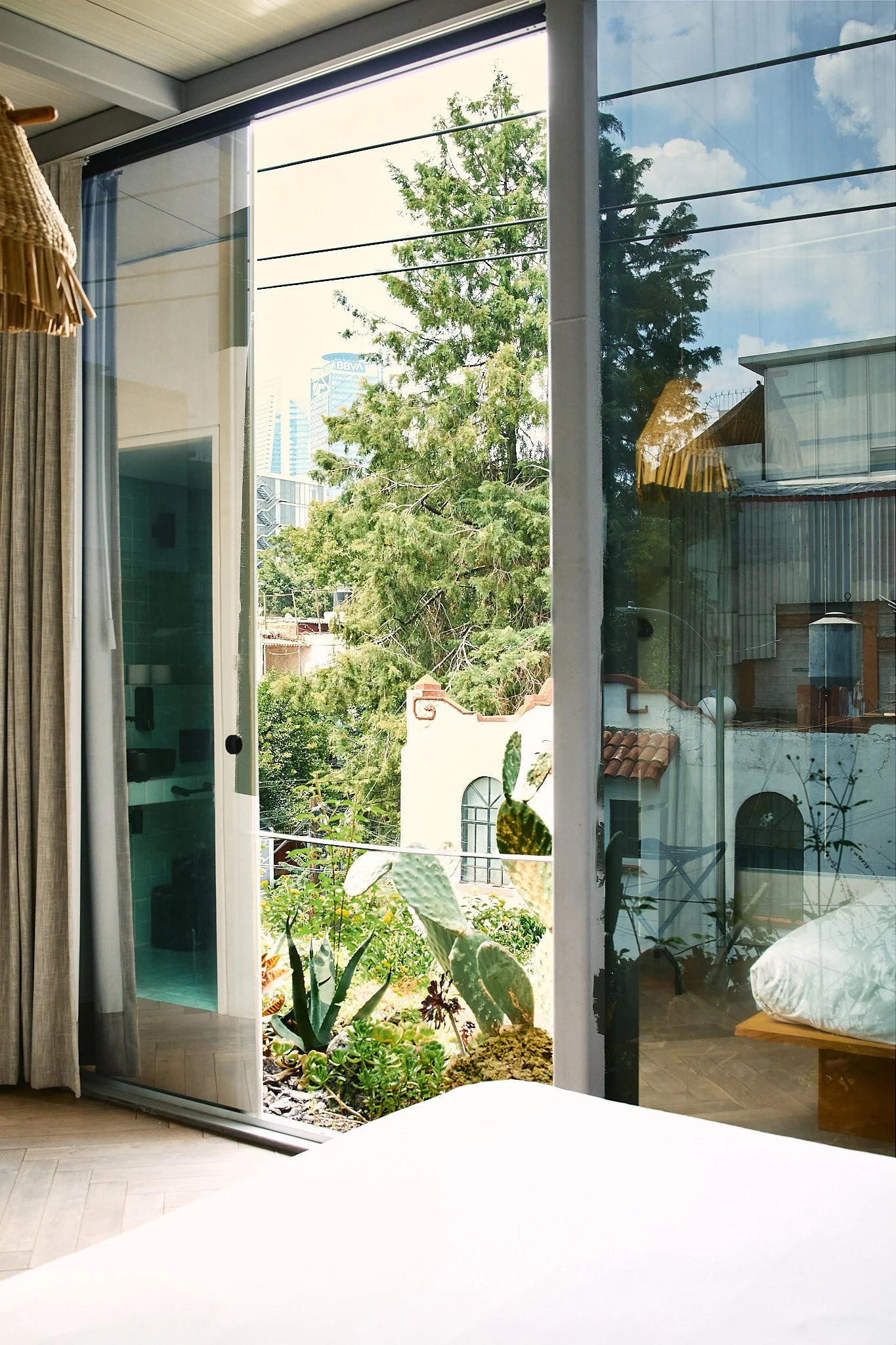 View through a large sliding glass door showing a small garden with succulents and an aloe plant, with a cityscape in the background including a tall building with the BBVA logo.