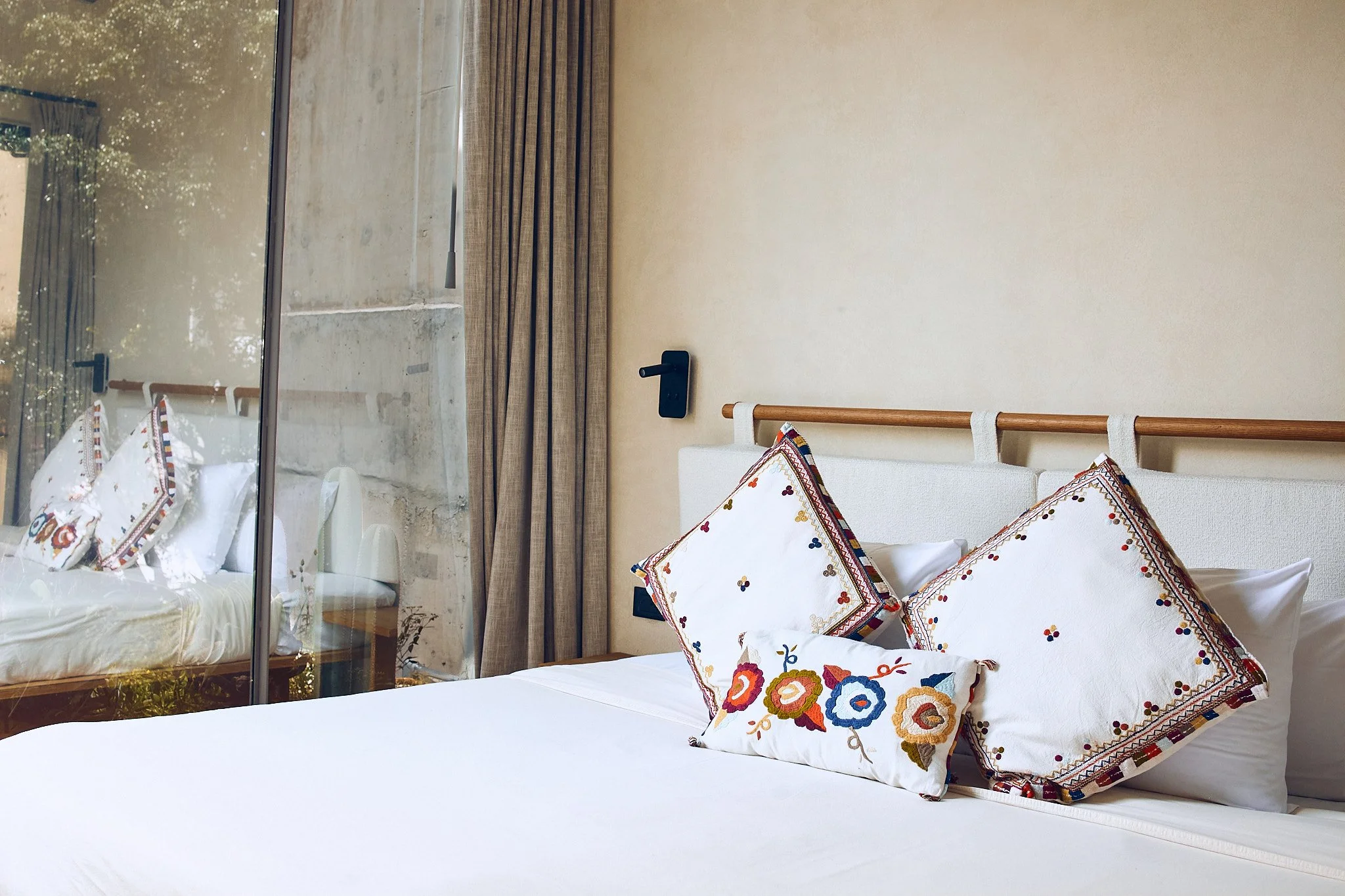 A bedroom with a bed that has embroidered pillows, a beige wall, a black wall-mounted light switch, and a large window with beige curtains reflecting sunlight.