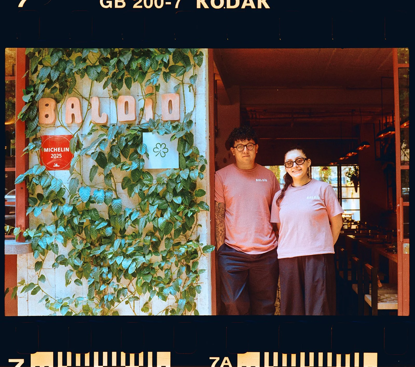 Two employees standing at the entrance of a restaurant named 'Baloii', with green leafy plants and a Michelin 2025 sticker on the wall.