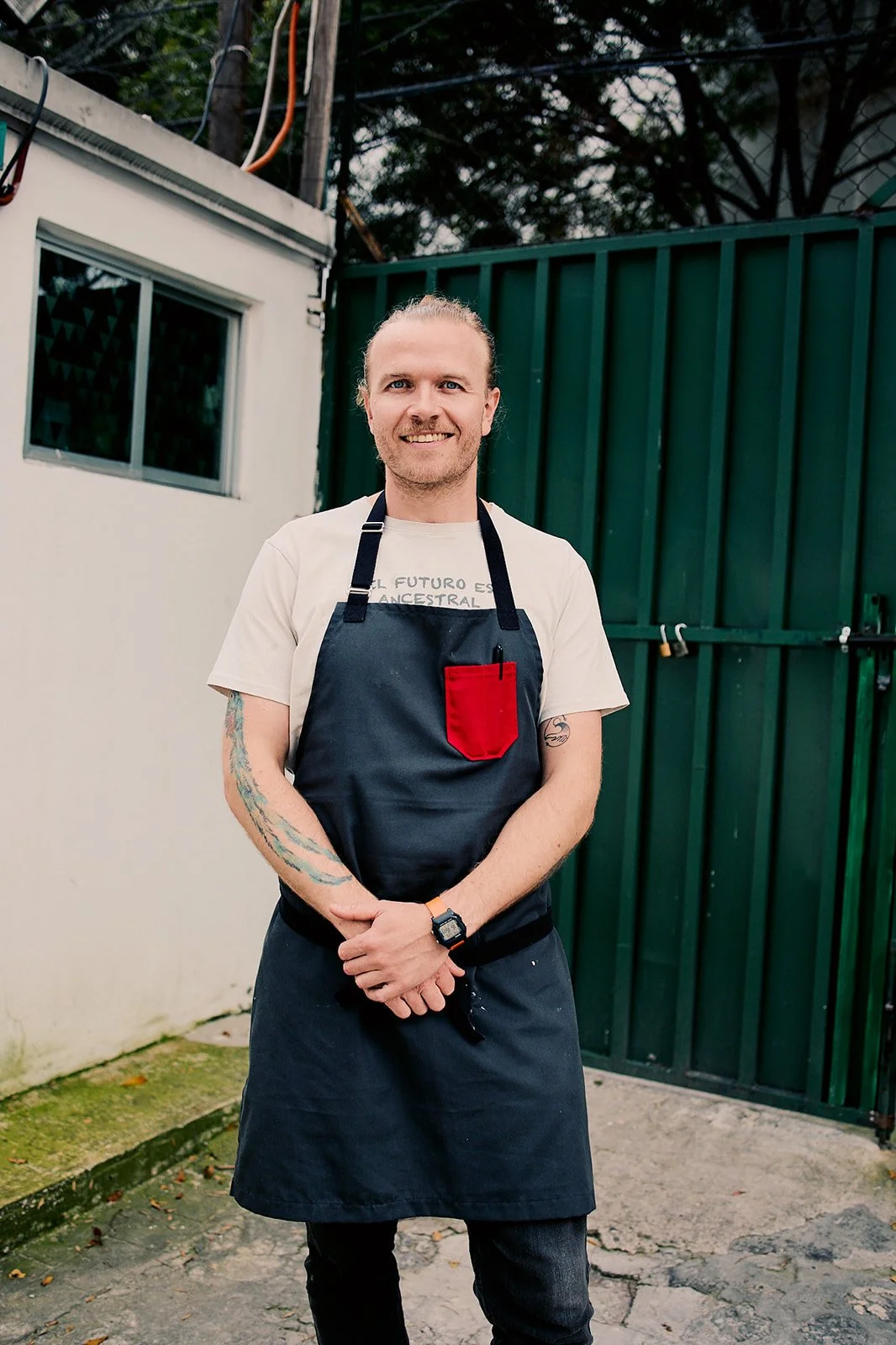 A man with long hair and tattoos wearing a white t-shirt, a black apron with a red pocket, and a wristwatch, standing outdoors in front of a green metal fence and building wall.