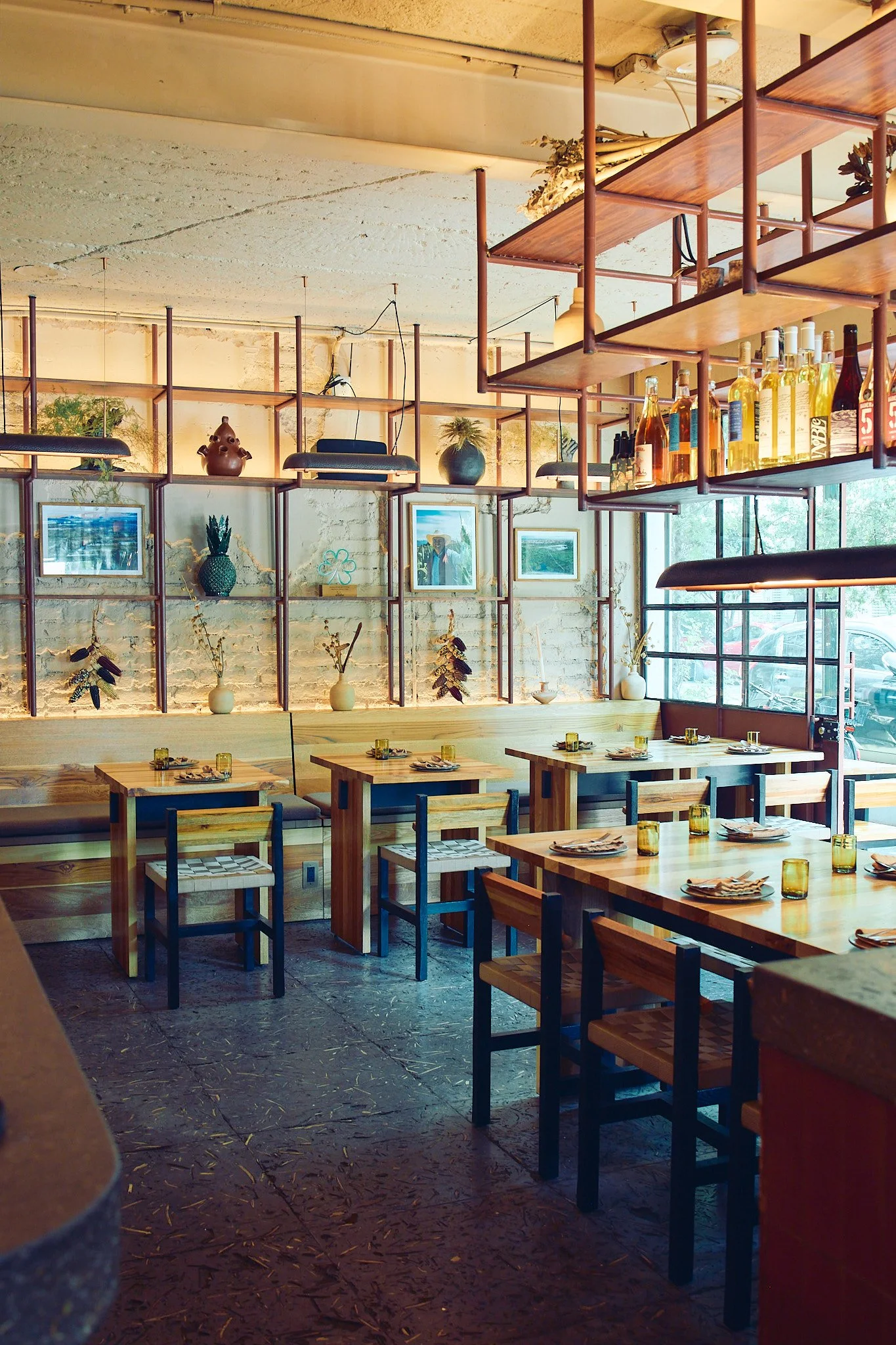 The interior of a restaurant with wooden tables set for dining, decorated with placemats, forks, and glasses. There are open shelving units decorated with plants, art, and bottles, against a white brick wall. Large windows let in natural light.