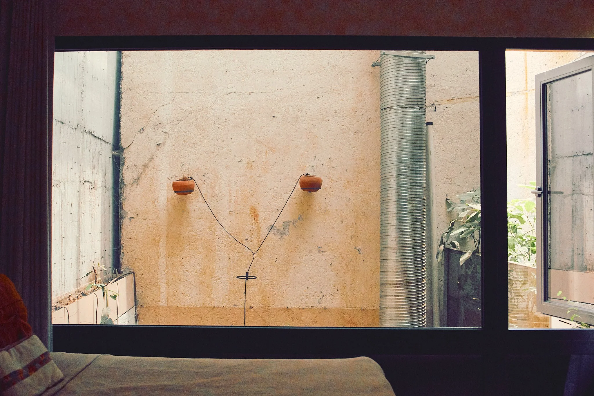 View from inside a room through an open window, showing a beige textured wall, a large silver ventilation pipe, and a small plant outside.