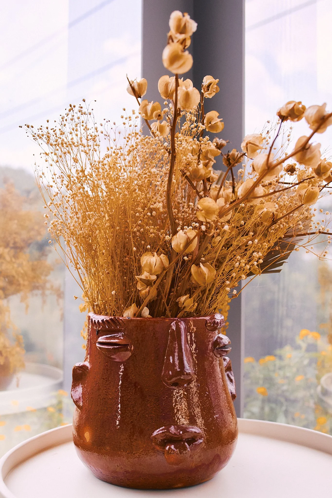 A brown ceramic vase with a face design, filled with dried flowers, placed on a white tray in front of a window.