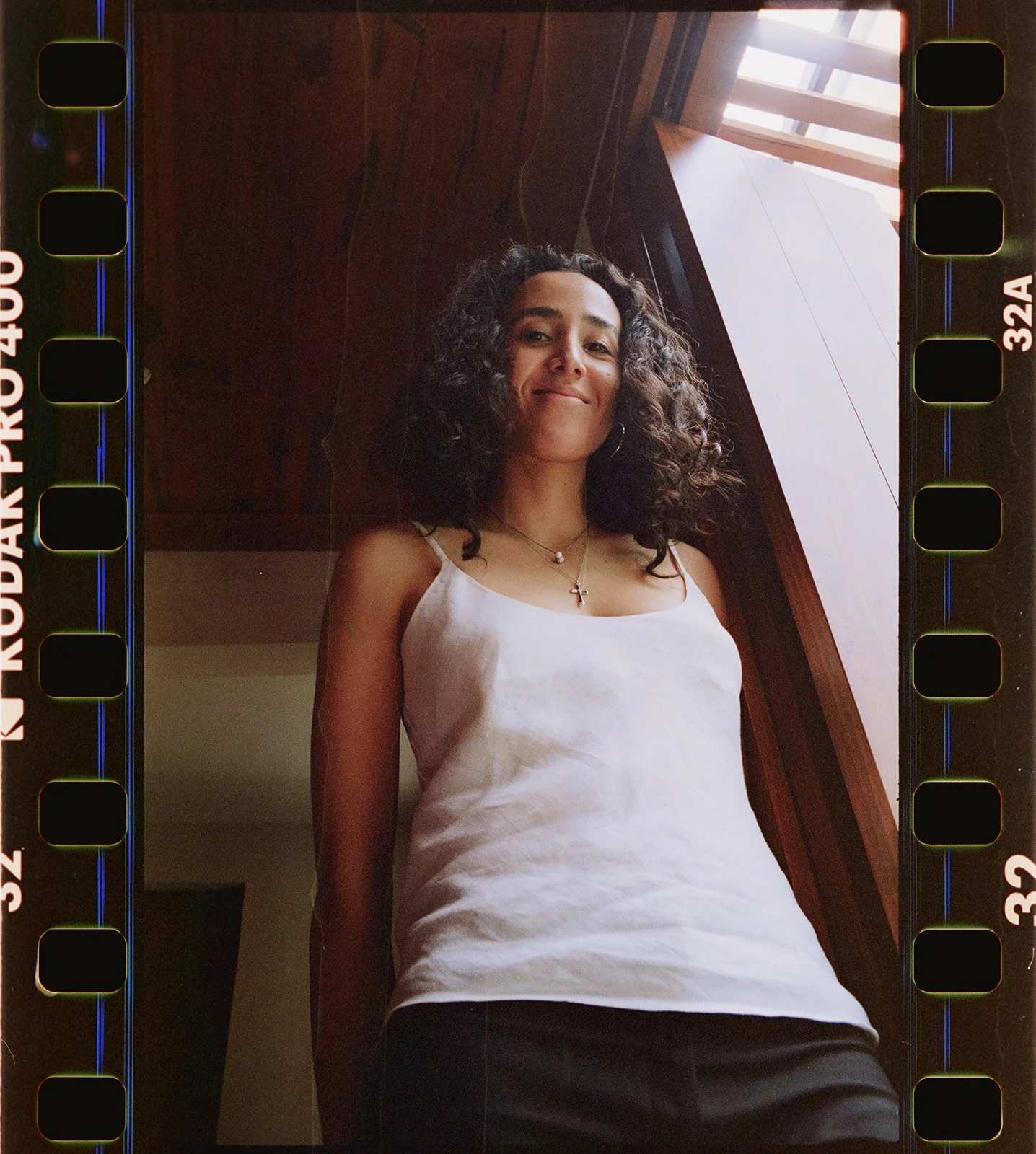 A woman with curly hair smiling and looking at the camera, wearing a white tank top and jewelry, standing indoors near a wooden ceiling and a skylight.