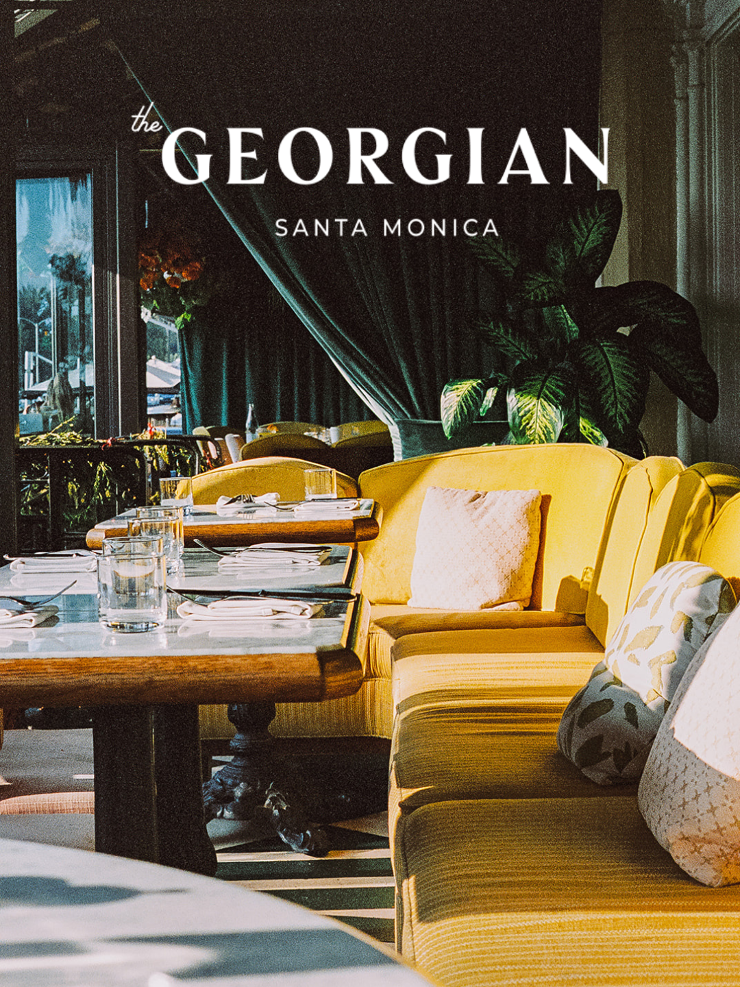 Interior of a restaurant with yellow upholstered booths, set tables with glasses and napkins, large green plant, dark curtains, and a window with outdoor view. Overlaid with text: 'the GORGIAN SANTA MONICA'.