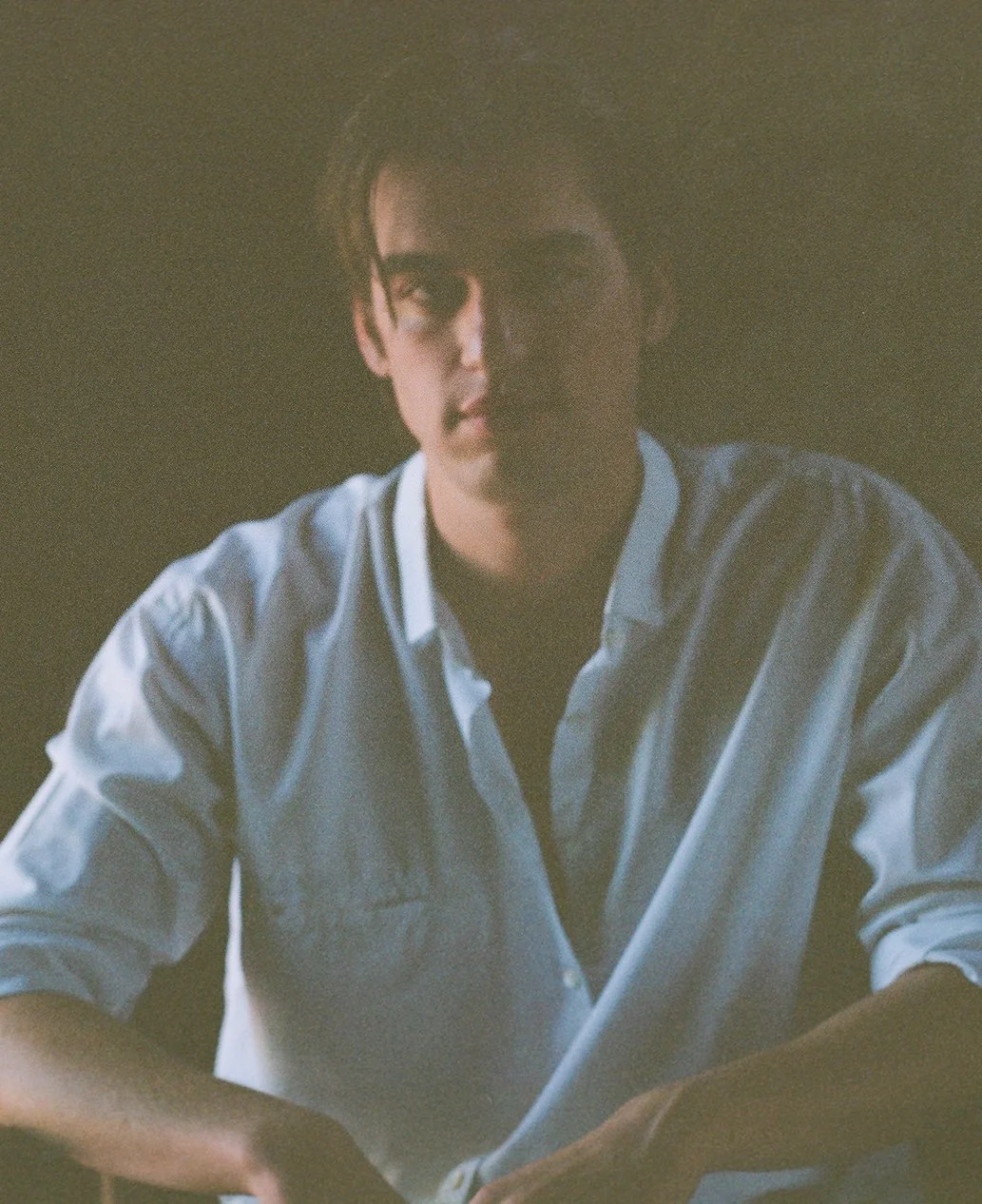 A young man with dark hair and glasses, wearing a light blue button-up shirt, sitting in a dimly lit room.