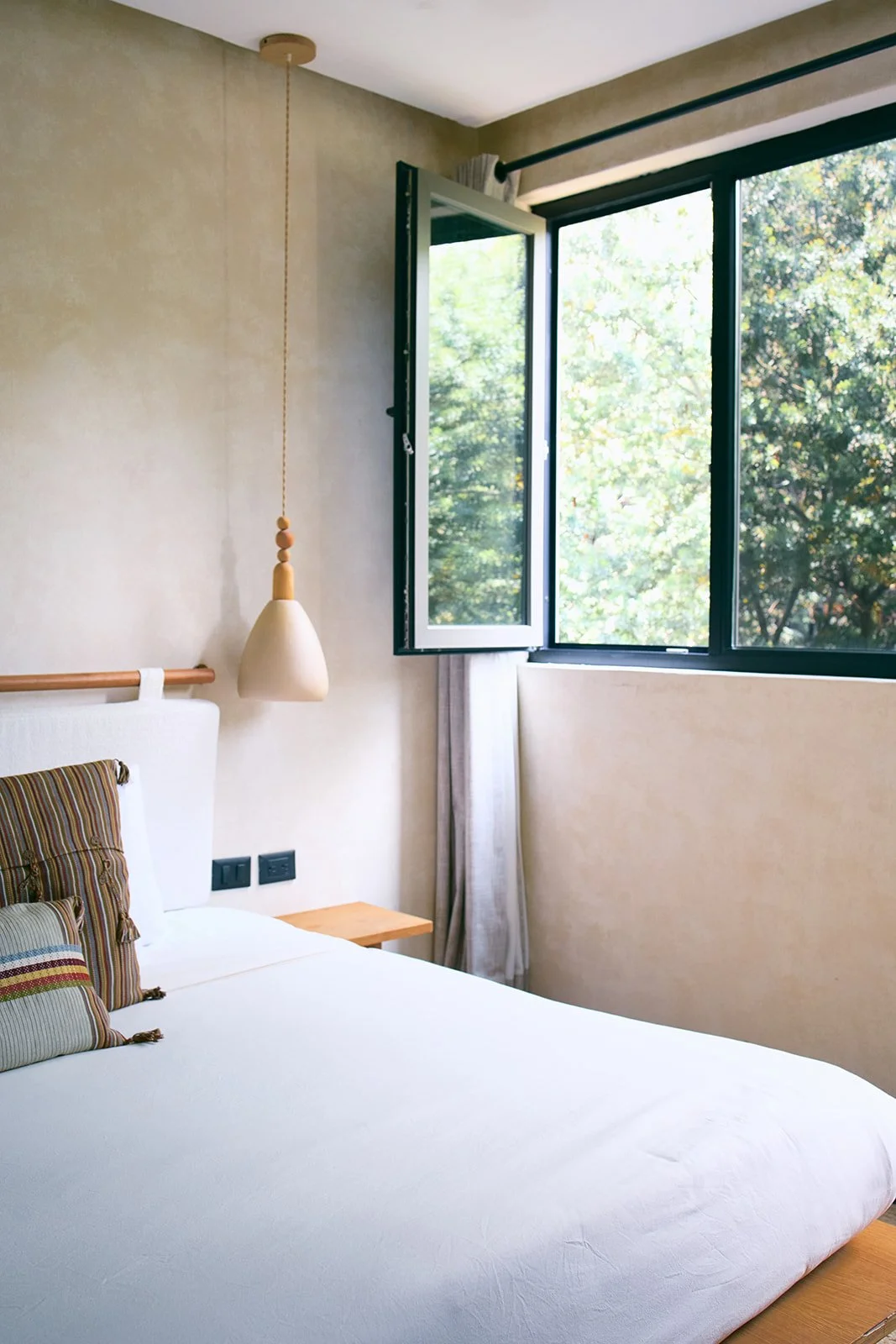 A bedroom with an open window showing green trees outside, a white bed with striped pillows, a bedside table, and a hanging pendant lamp.