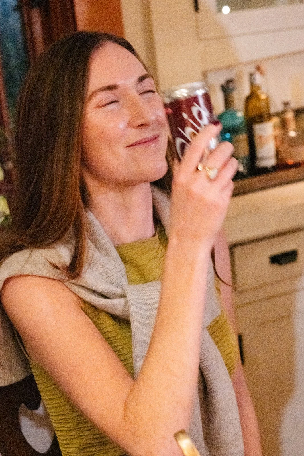 A woman with long brown hair smiling with her eyes closed, holding a can of Coke close to her face in a kitchen setting.