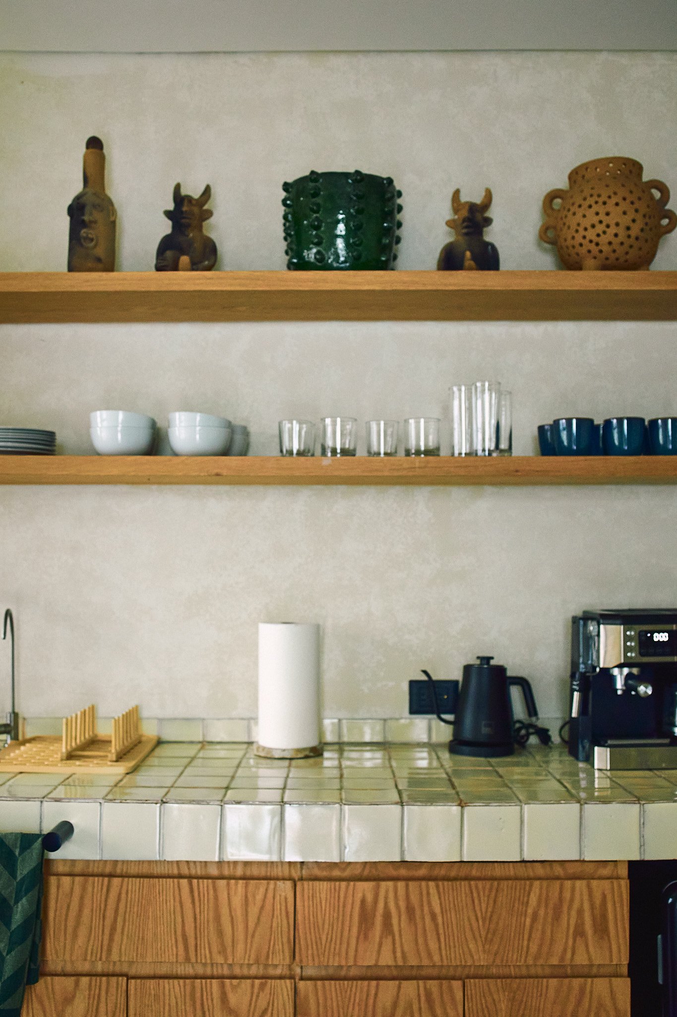Kitchen counter with white tile surface, paper towel roll, black electric kettle, coffee machine, white bowls, glass cups, wooden dish rack, and wooden cabinets, with a wooden shelf above holding decorative pottery.