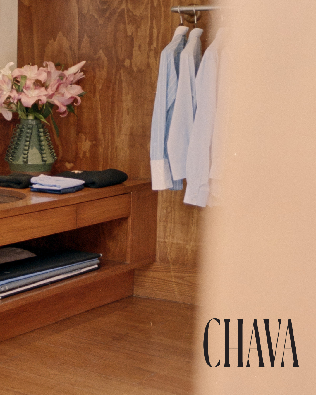 A wooden closet with hanging white dress shirts and pants, a bouquet of pink lilies in a green vase, and neatly folded clothes on a shelf, with the brand name 'CHAWA' in stylized black font.
