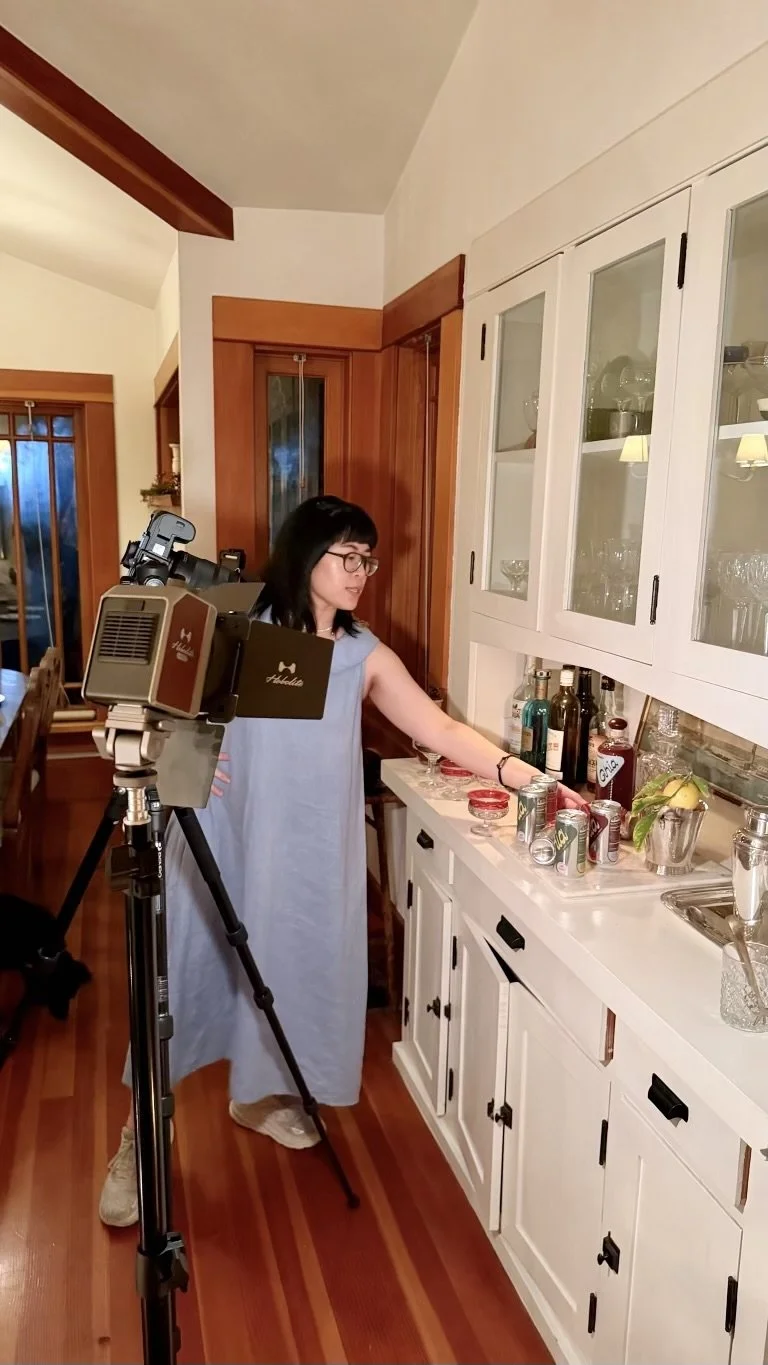 A woman with glasses and black hair, wearing a light blue dress, standing in a kitchen next to a white cabinet with glass doors. She is reaching for items on the counter, which has bottles, cans, and jars. There is a camera on a tripod in front of her, capturing her activity.