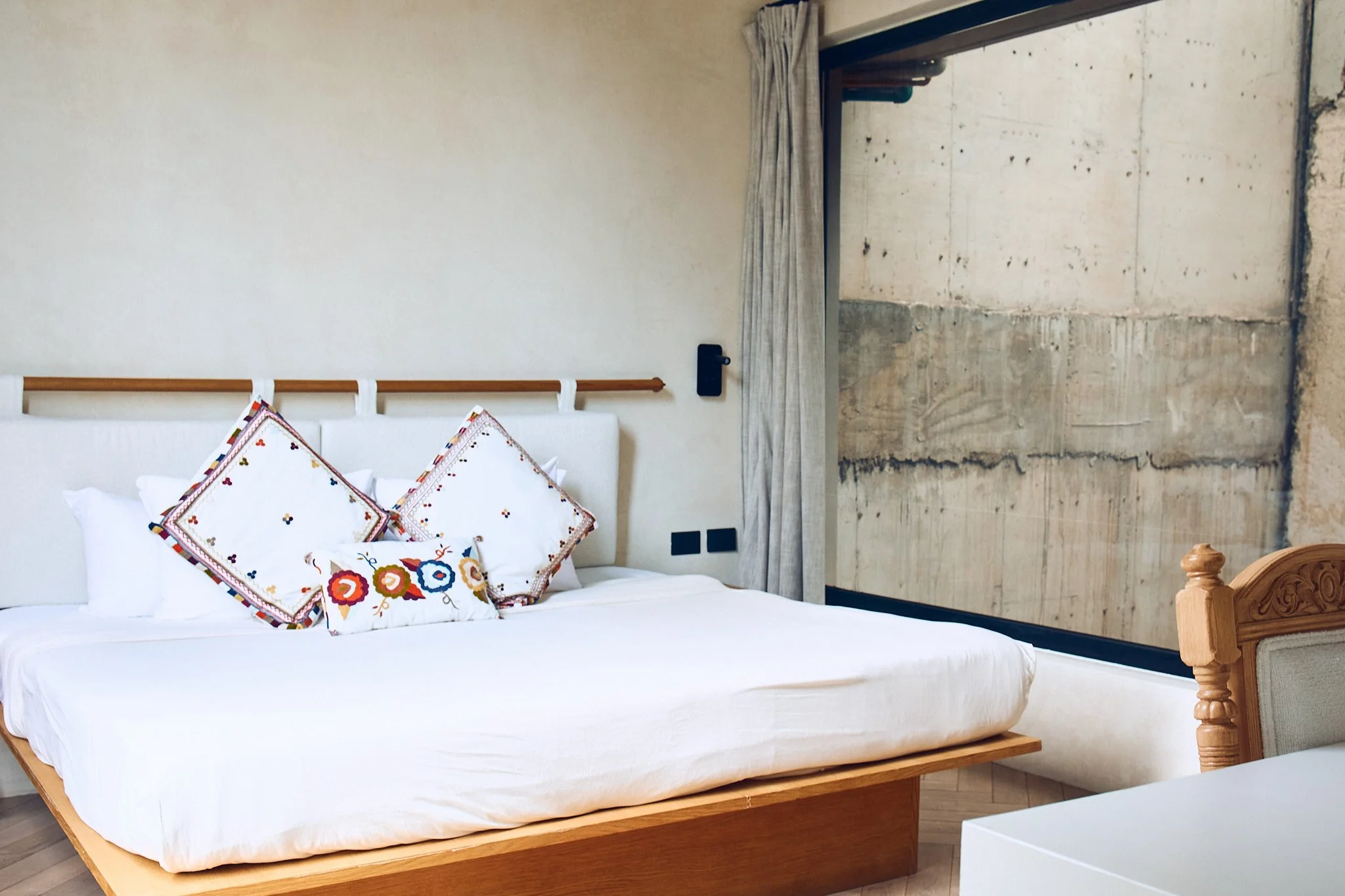 A bedroom with a wooden bed frame, white bedding with colorful embroidered pillows, and a concrete wall with a sliding glass door covered by beige curtains.