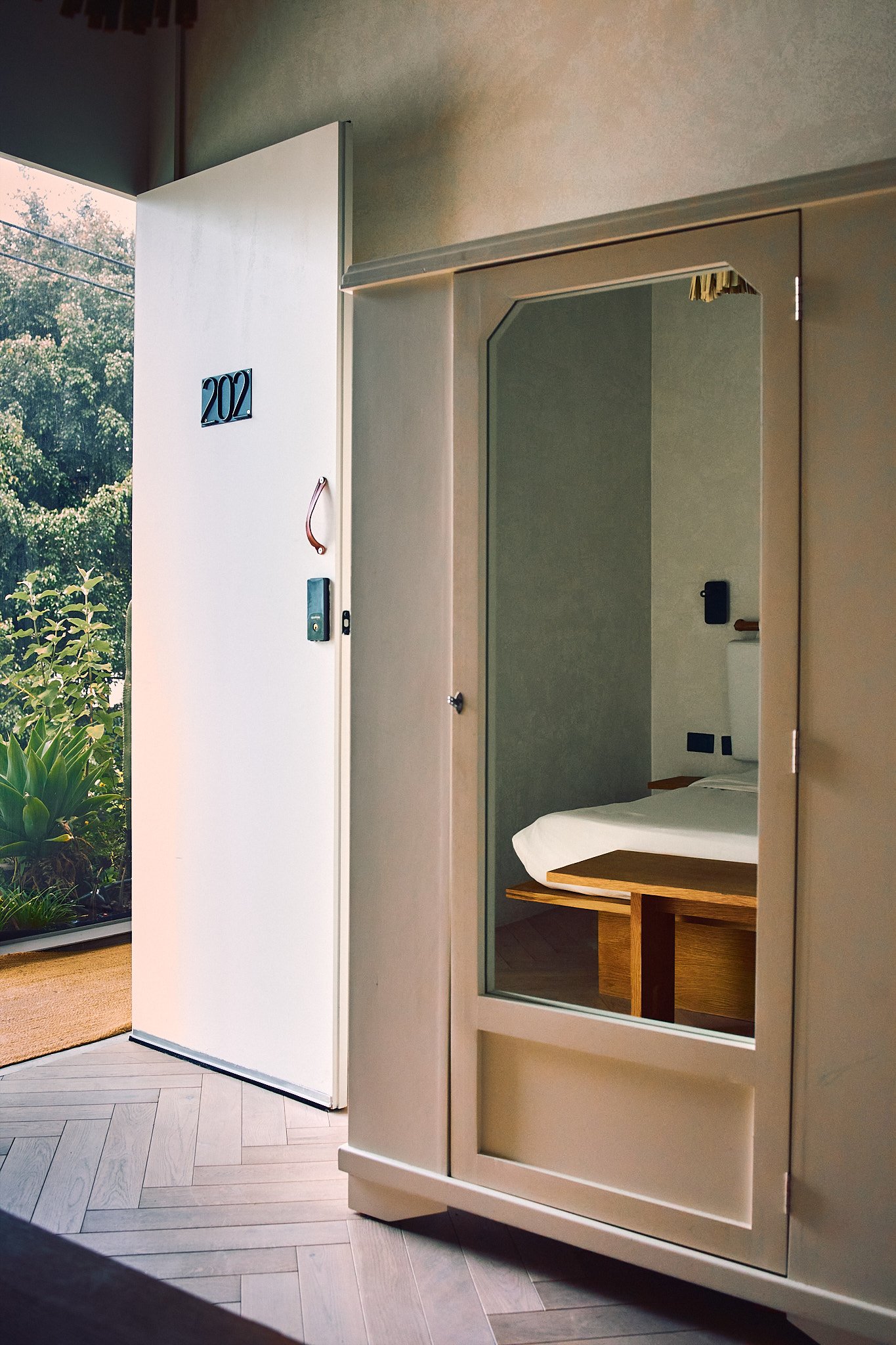 Open hotel room door revealing a bed, a wooden side table, and a mirror, with greenery visible outside.