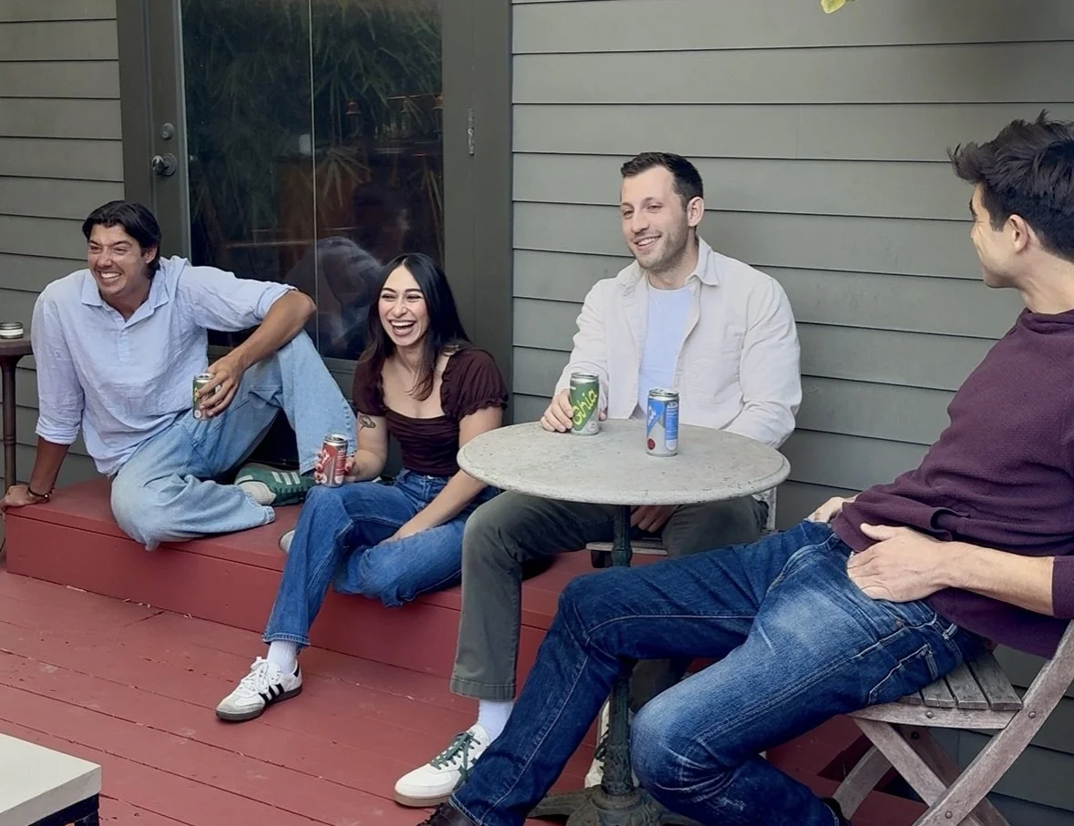 Four young adults sitting on a porch, laughing and chatting, with drinks in their hands. One person is sitting on an elevated platform, and the others are seated at a small round table and on chairs.