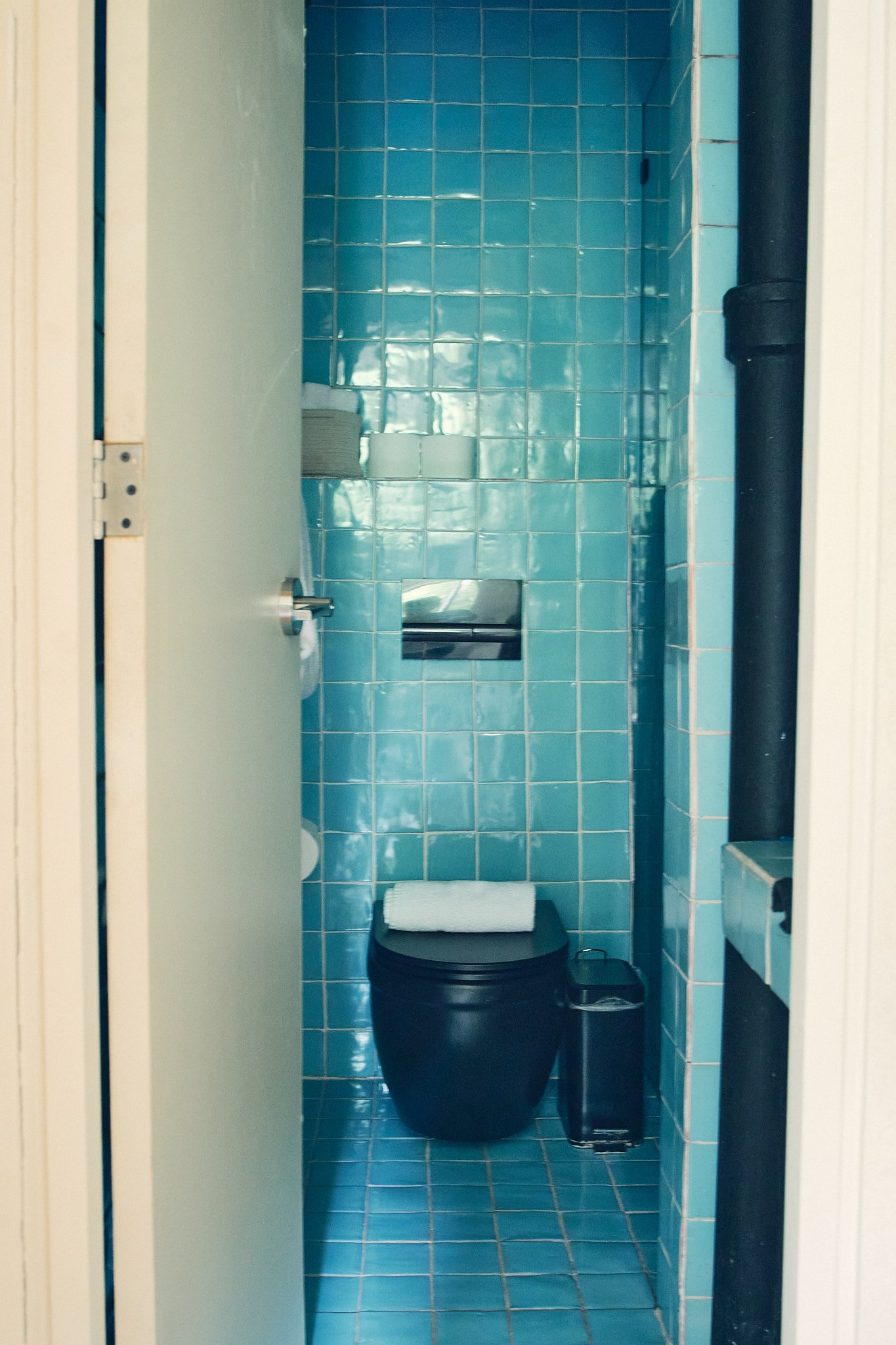 A small bathroom with blue tiled walls and floor, with a black toilet, a white roll of toilet paper on a paper holder, and a small black trash bin.