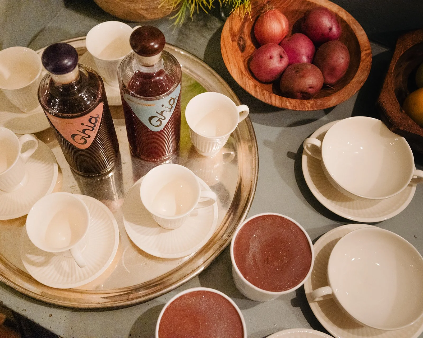 Table setting with coffee cups, two bottles labeled 'Chia', bowls of fruit including potatoes and yellow fruits, and glasses of red juice.