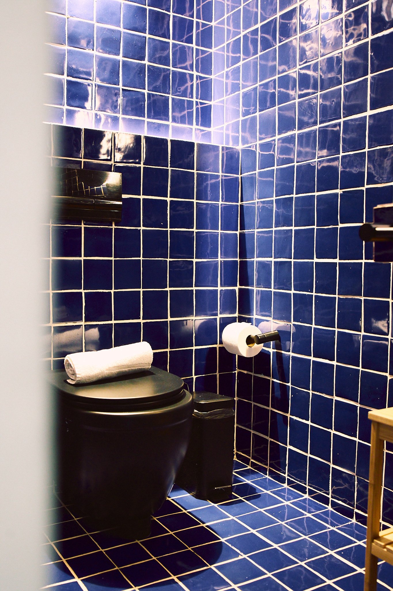 A small bathroom with blue tiled walls and floor, a black toilet with a white towel on top, a mirror, toilet paper on a holder, and a wooden shelf.