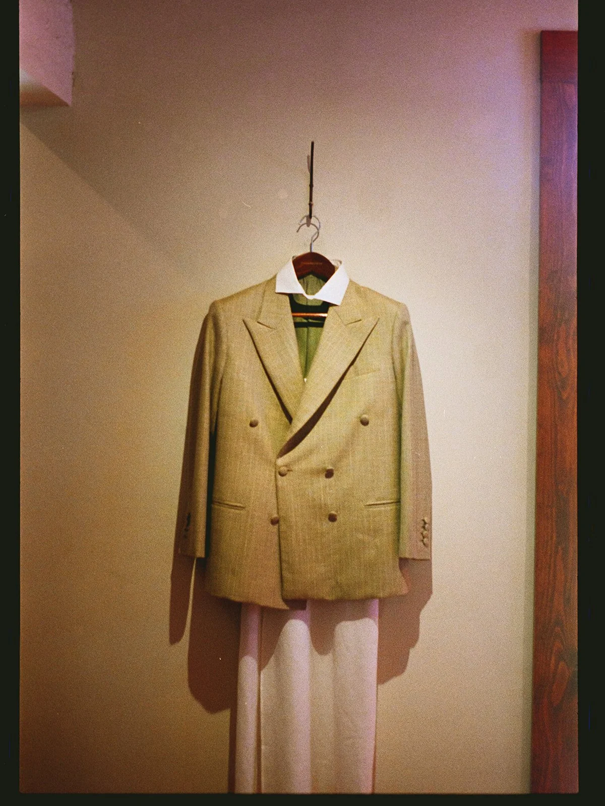A beige double-breasted blazer hanging on a wooden hanger with a white collar shirt underneath, displayed against a plain wall.