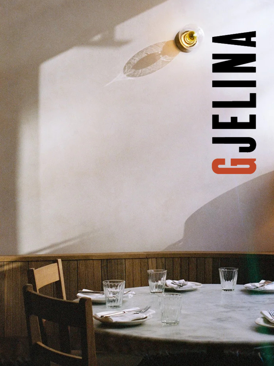 Empty restaurant table with glasses and napkins, beige wall with a projection of a light bulb and shadow, and the word "GELINA" in black and orange text.