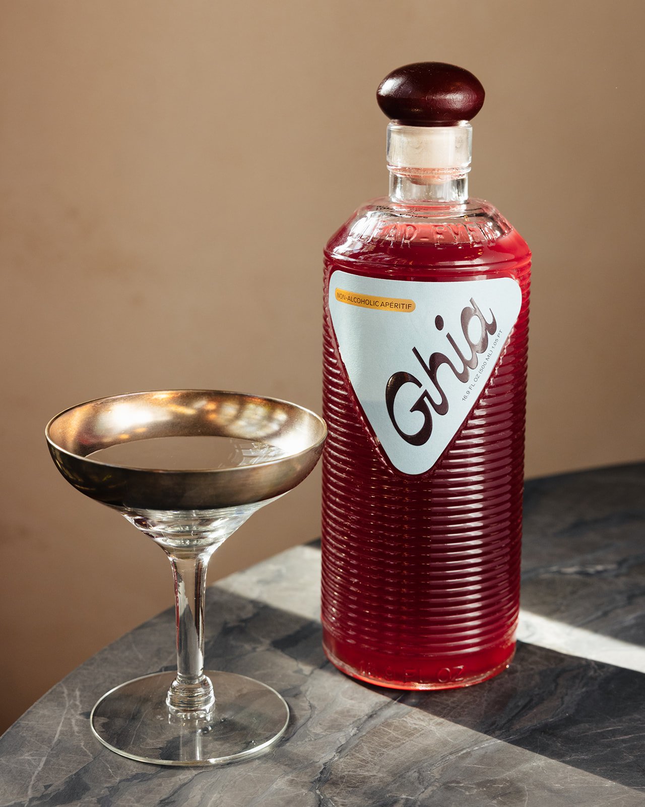 A cocktail glass with a dark liquid next to a tall bottle of red Ghia non-alcoholic aperitif on a marble table.