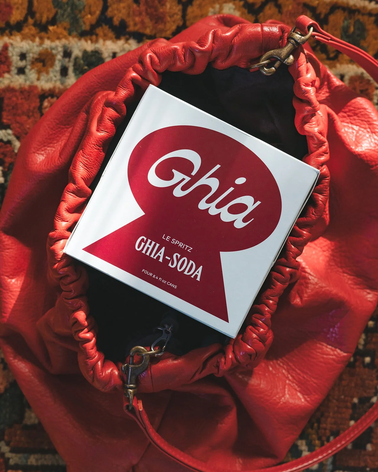 A red leather bag with a round, gathered opening and brass chain detail, inside of which is a box labeled "Ghia" containing four cans of Ghia-Soda.