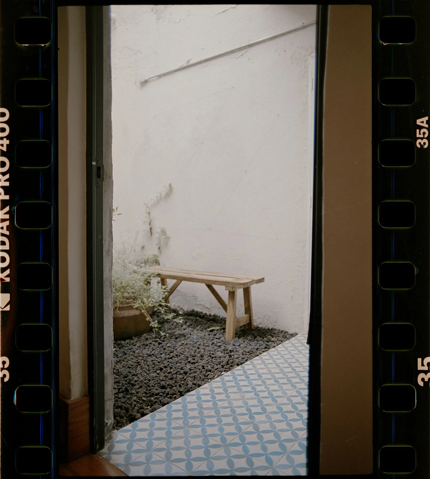 View of a small outdoor space through a doorway, featuring a white textured wall, a wooden bench, a potted plant, and gravel ground, with a patterned tile flooring near the entrance.