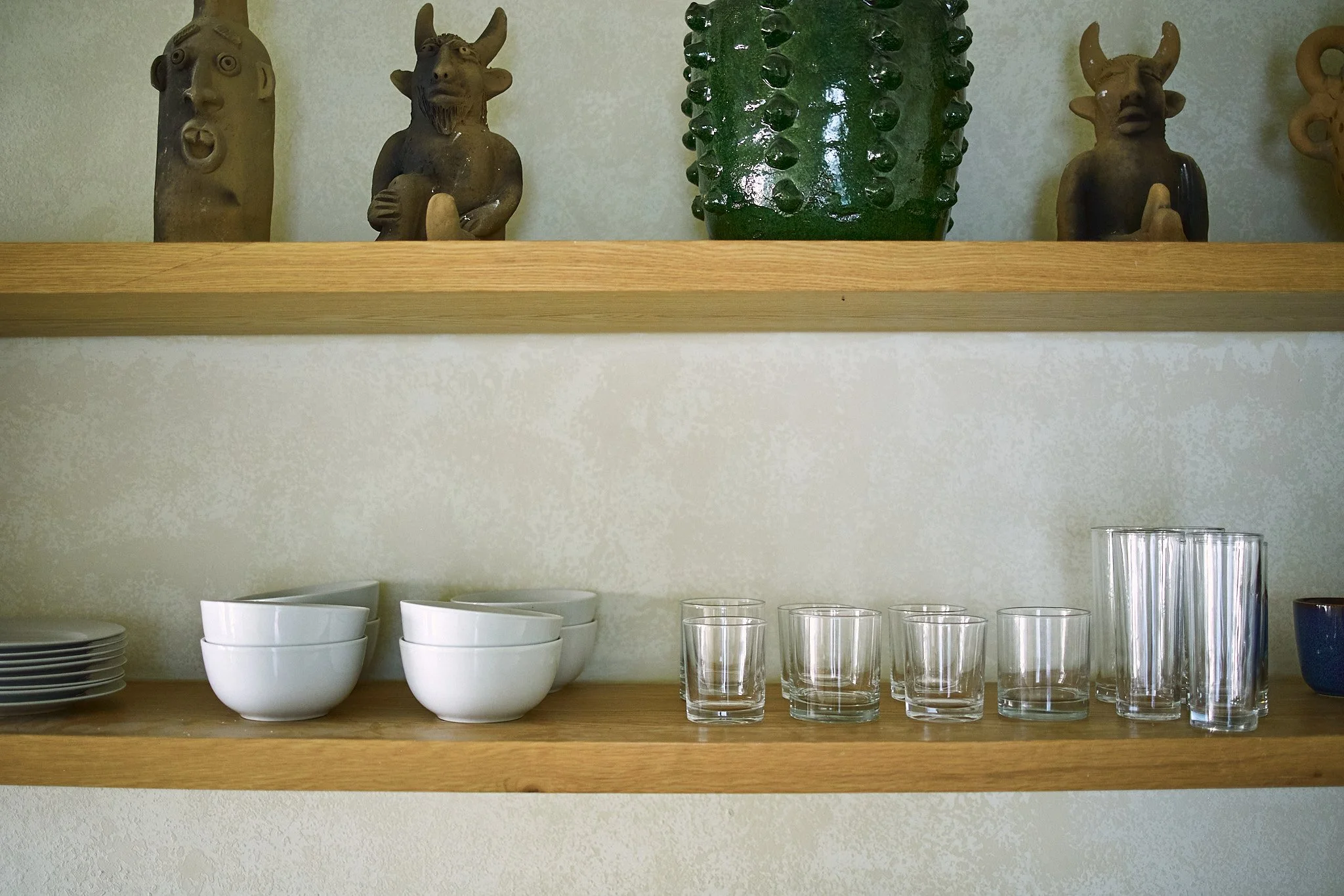 Wooden kitchen shelf holding decorative ceramic figures and green glass vase, with white bowls, plates, and clear drinking glasses below.