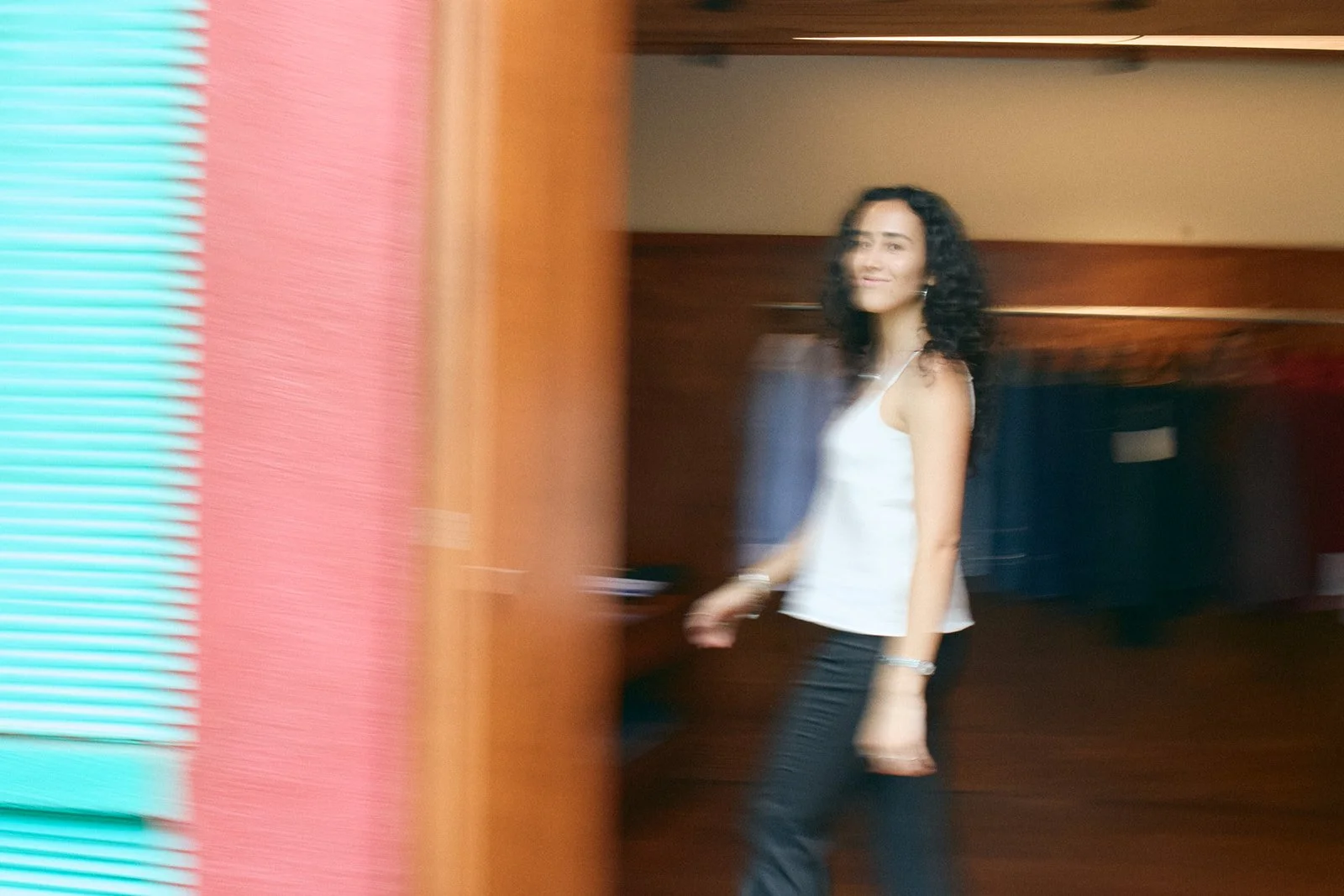 A woman with curly hair wearing a white tank top and black pants, walking indoors with a blurred background.