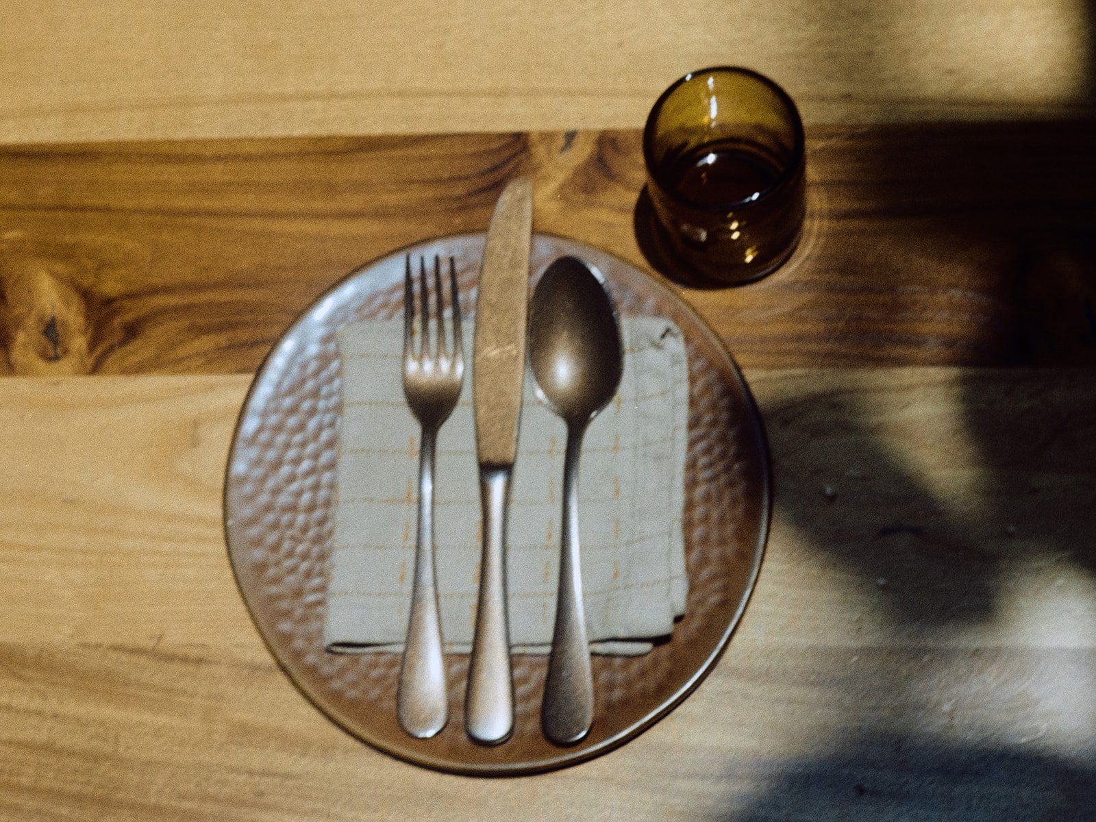 A wooden table with a glass of dark-colored beverage, a small plate with a fork, knife, and spoon resting on a napkin.