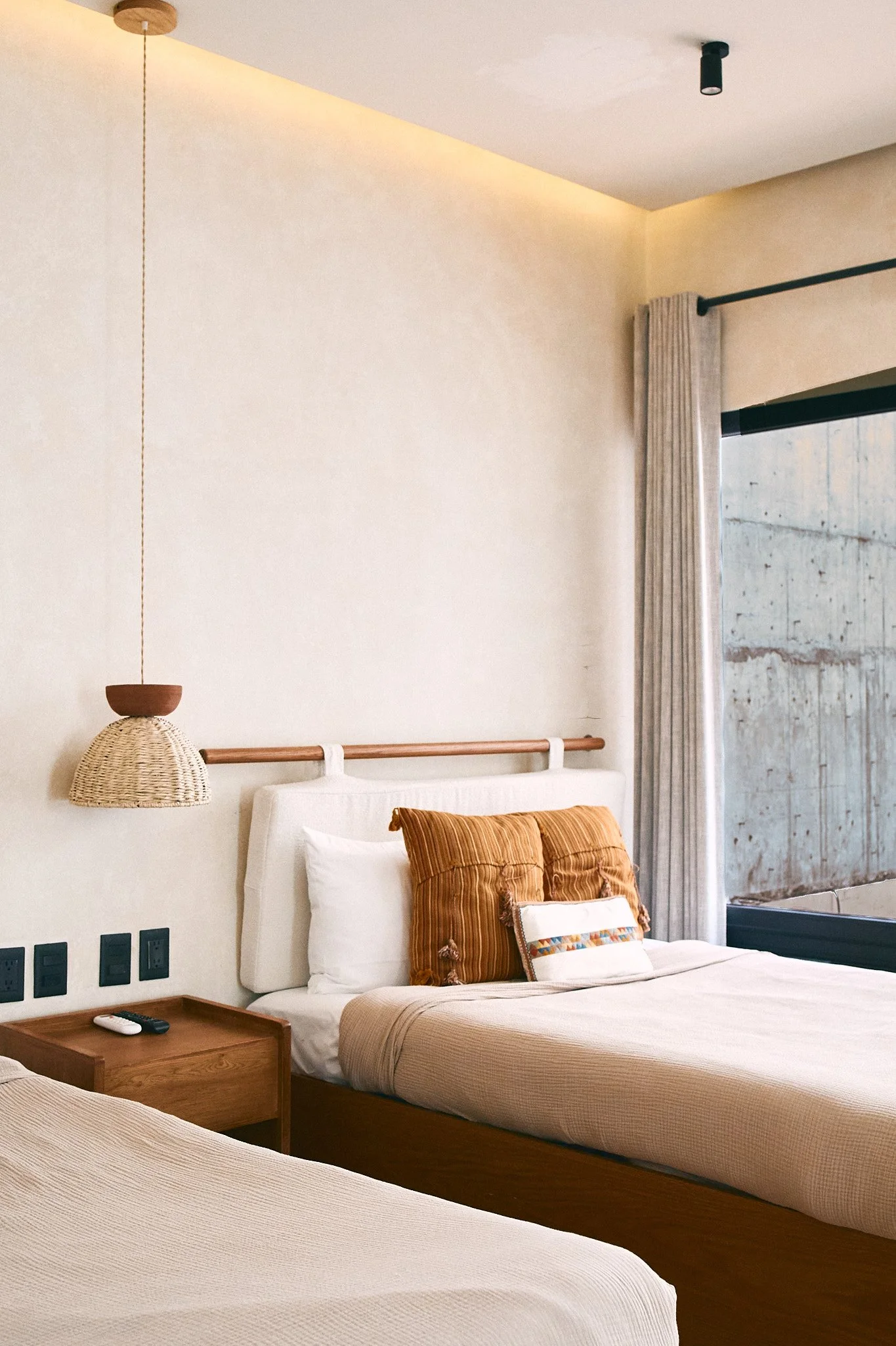 A bedroom with beige walls, a bed with white linens, orange and striped pillows, a wooden nightstand with a remote, a woven pendant lamp, beige curtains, and a window with a concrete wall outside.
