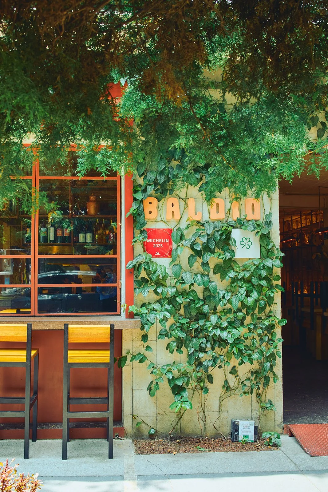 Exterior of a restaurant named 'Balido' with green vines and leafy plants growing on the wall. There is a red Michelin 2025 badge on the wall and a window showing bottles inside. Two yellow and black chairs are outside.