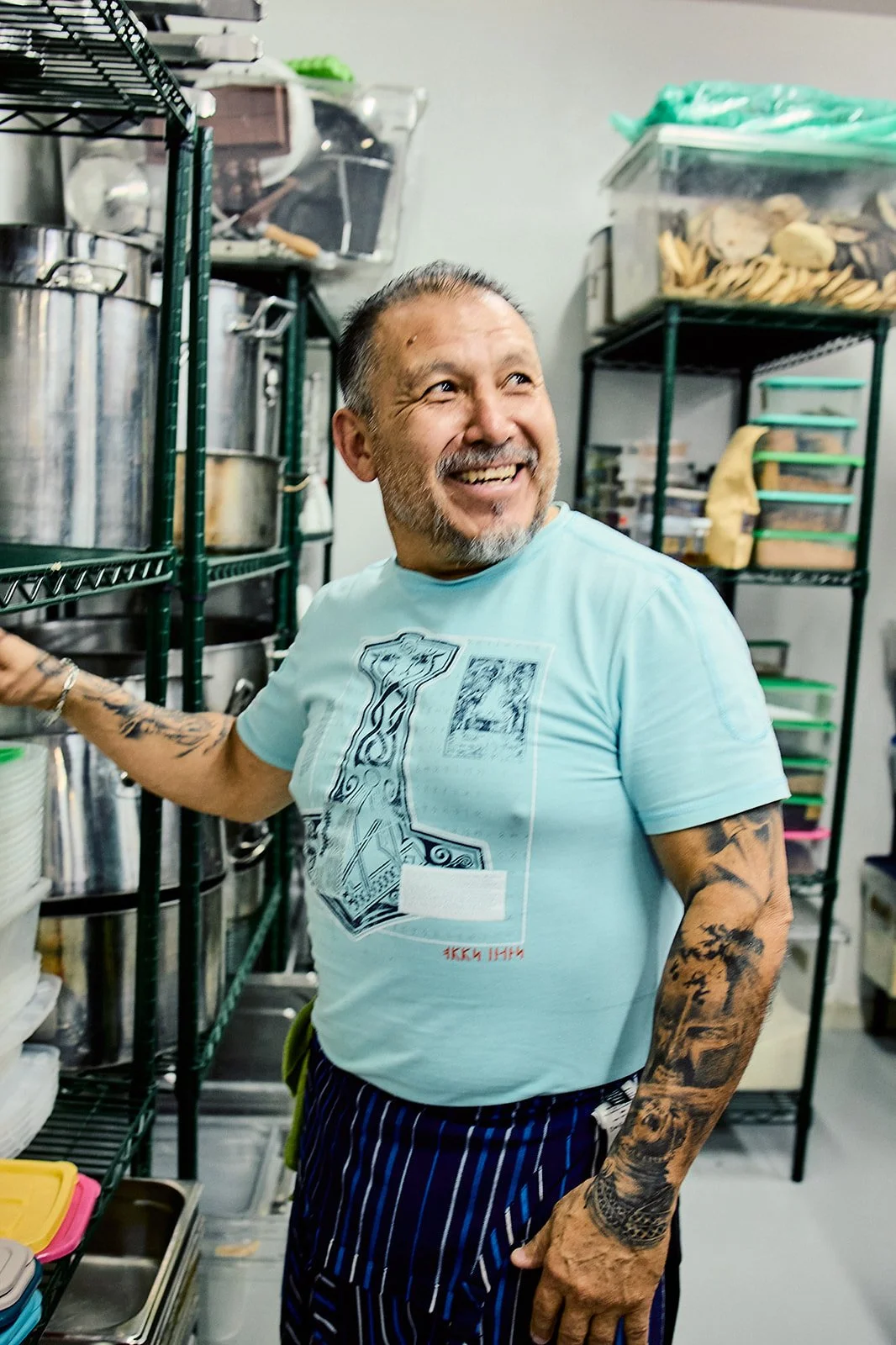 A smiling man with tattoos on his arms, wearing a light blue t-shirt with a graphic design, standing in a pantry or kitchen storage room filled with shelves of containers and food.