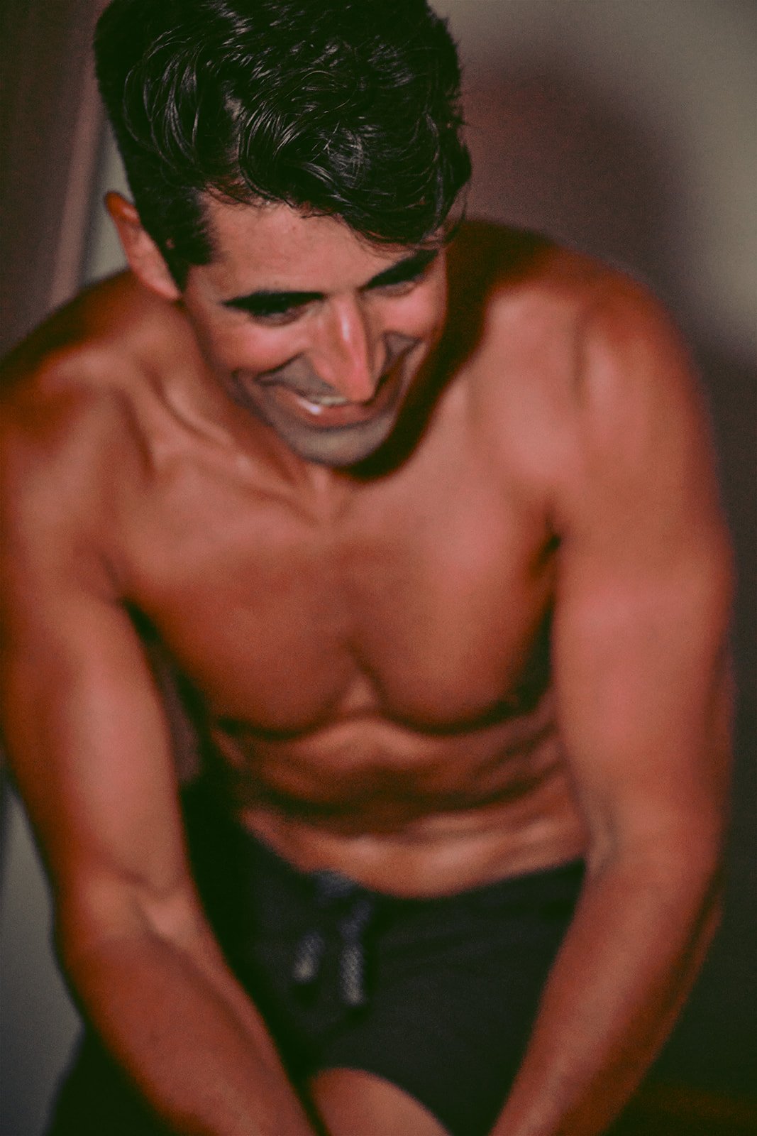 A shirtless young man with dark, curly hair smiling and looking downward, inside a room with a plain background.